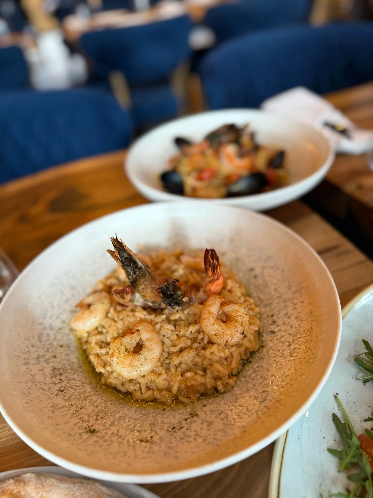 A bowl of seafood risotto with shrimp and prawns on a wooden table, with another dish featuring mussels and seafood pasta in the background.