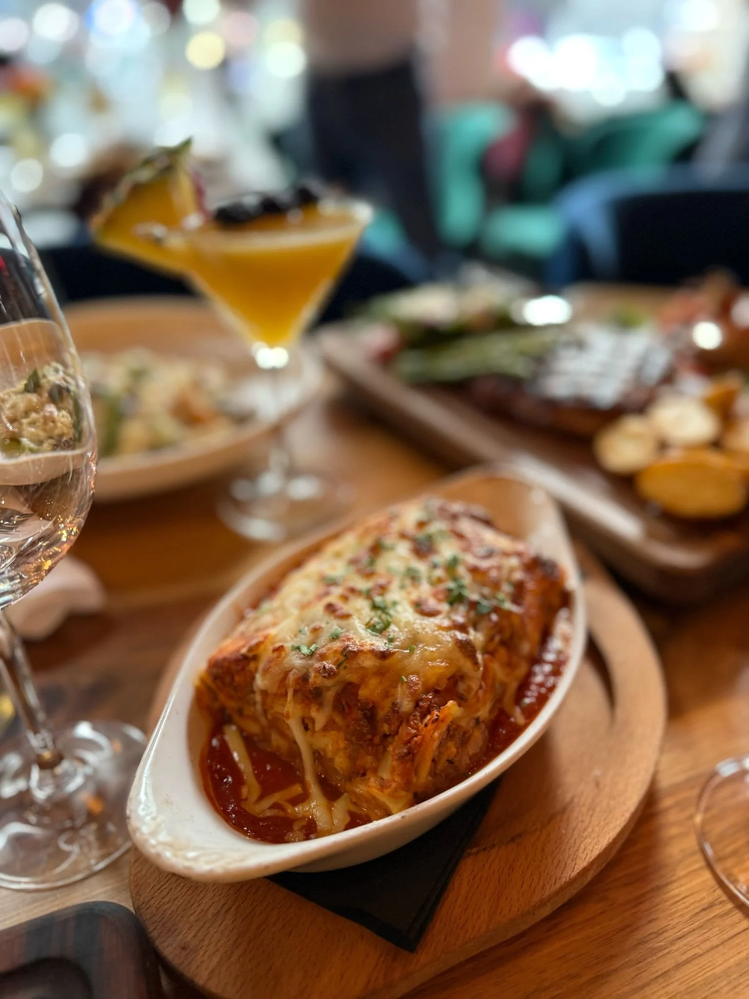 Close-up of a lasagna served in a white dish on a wooden board, with a glass of wine and a cocktail in the background, on a restaurant table.