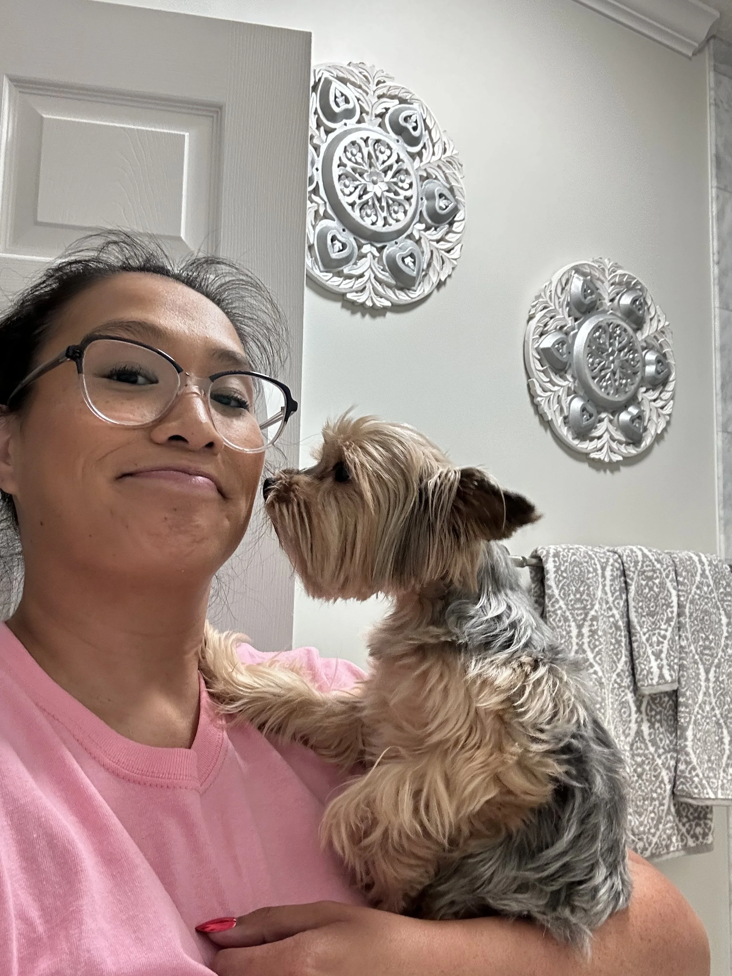 A woman holding a small Yorkshire Terrier dog in a bathroom, with decorative wall art and towels in the background.