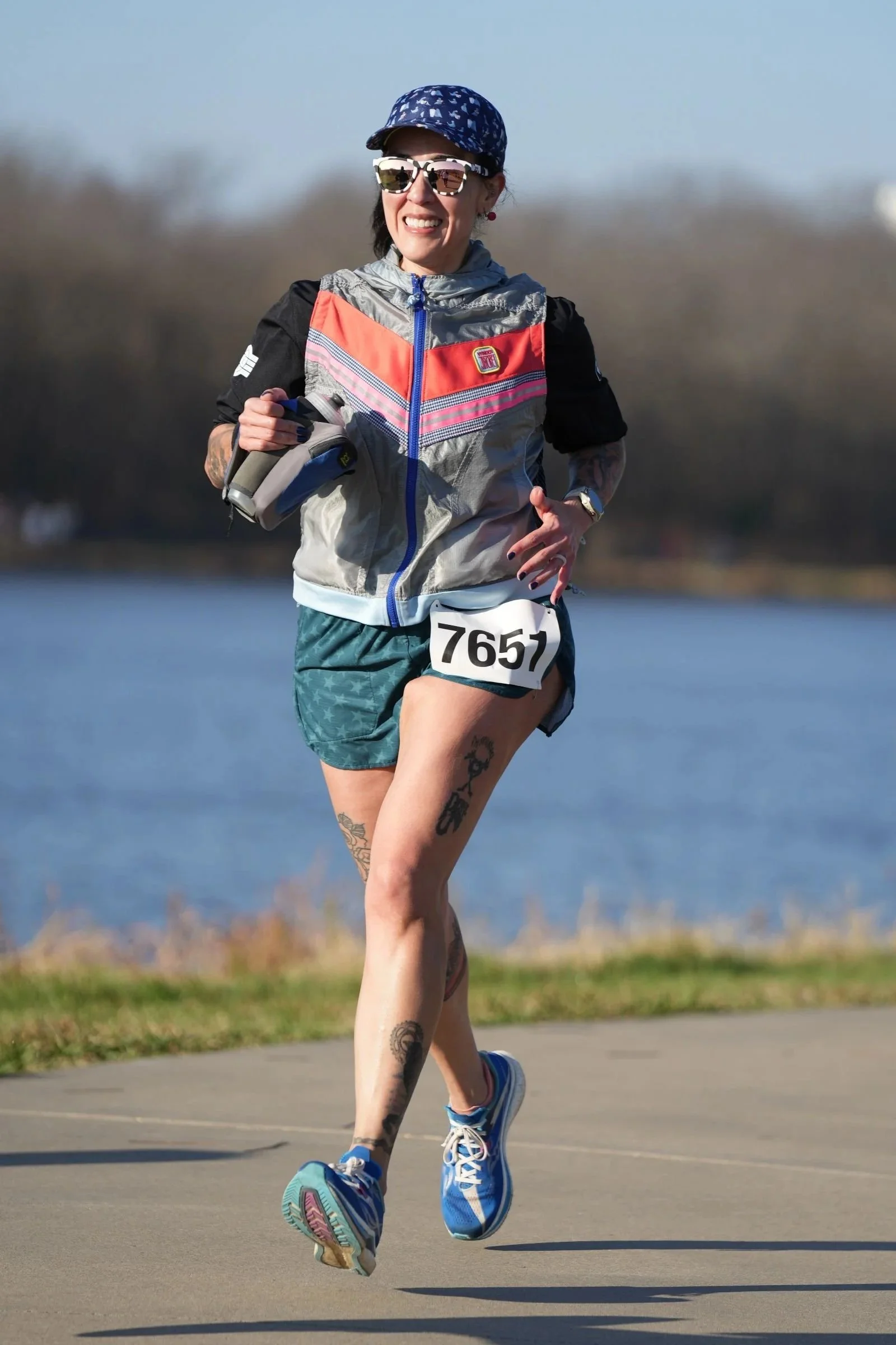 A woman runs outdoors near a body of water, wearing sunglasses, a cap, a colorful jacket, and shorts, with a race bib number 7657. She has tattoos on her legs and is smiling.