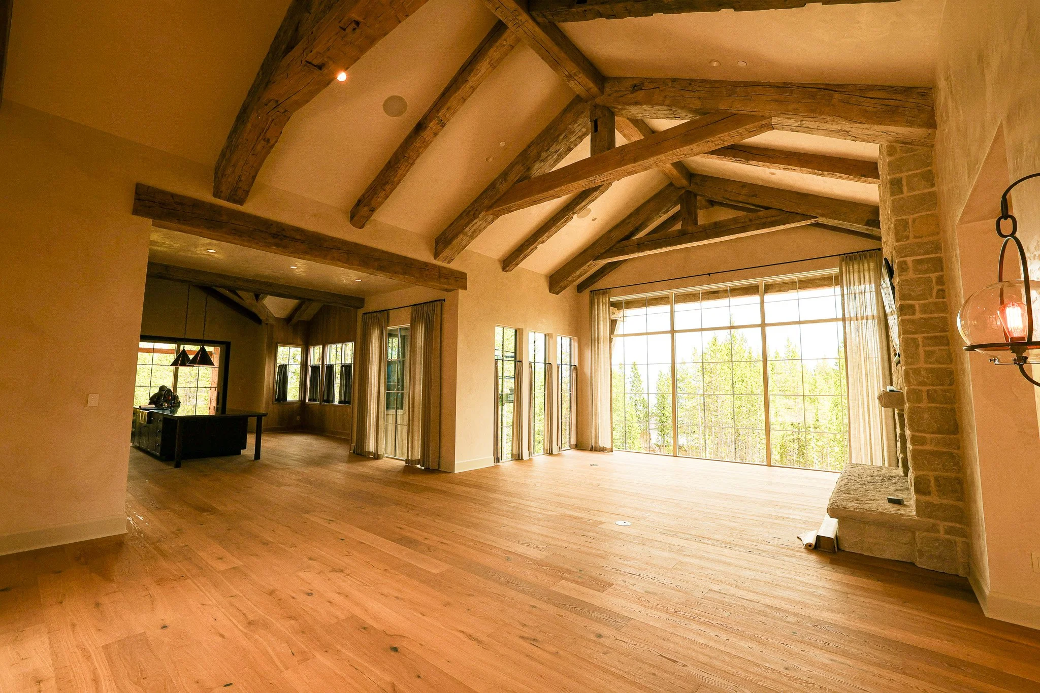 Spacious living room with large floor-to-ceiling windows showing greenery outside, exposed wooden beams on the ceiling, and a mix of brick and plaster walls, with hardwood floors and minimal furnishings.