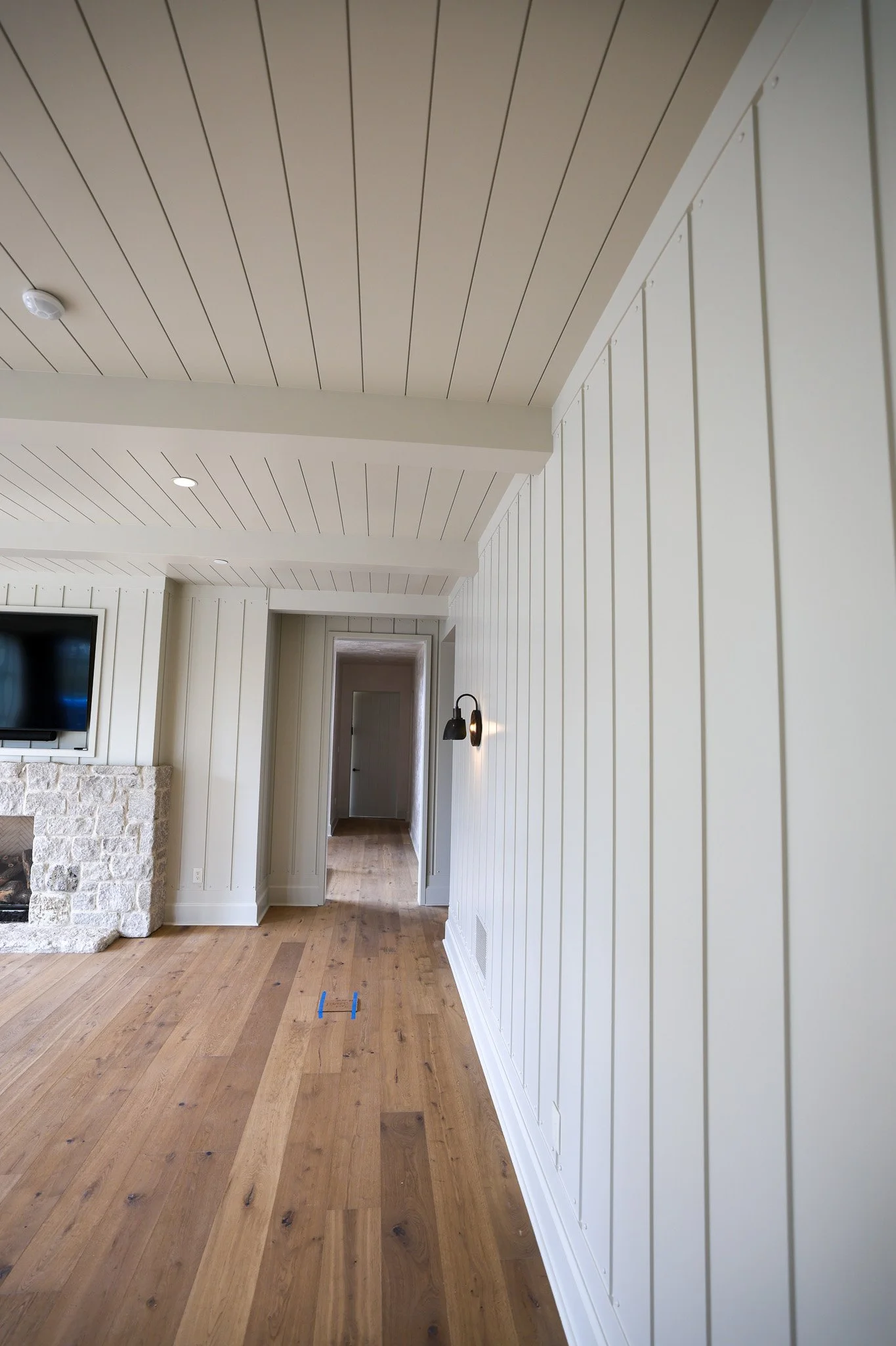 Interior view of a room with white vertical shiplap walls, a wood floor, a ceiling with white wooden panels, recessed lighting, wall-mounted fixtures, and a doorway leading to another room.