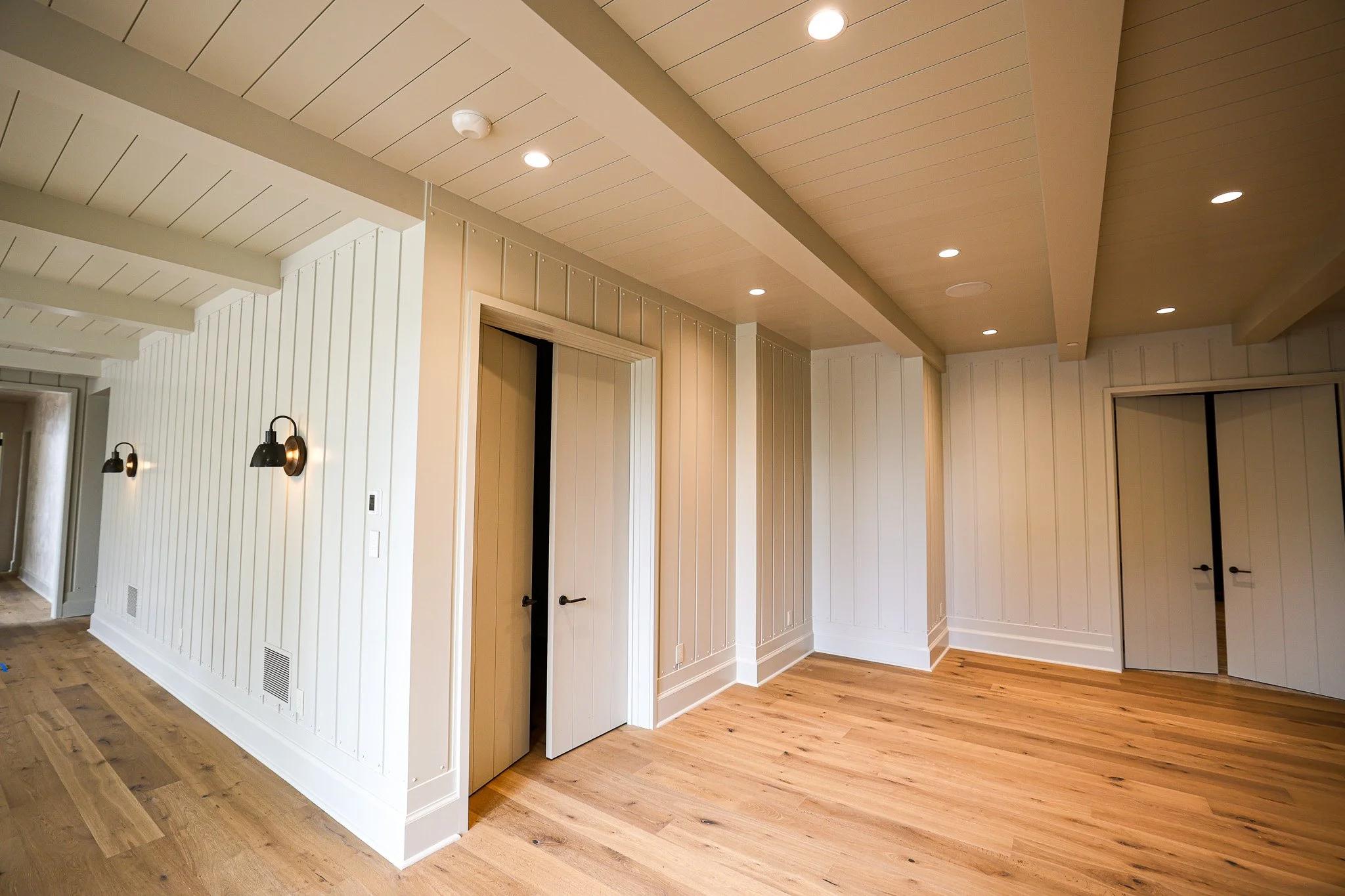 Interior view of a room with white shiplap walls, wooden flooring, and a ceiling with recessed lighting. There are two doors and wall-mounted light fixtures.