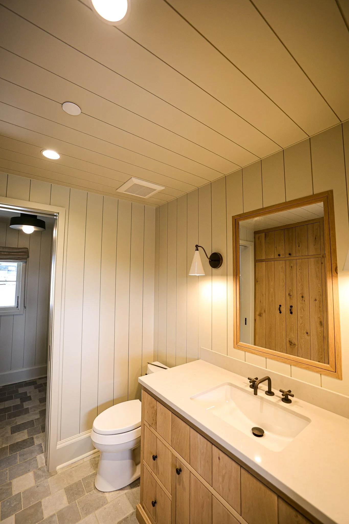 Bathroom with beige shiplap walls, wooden framed mirror, white sink with black fixtures, toilet, and a wall-mounted lamp. Visible in the background is part of a room with window and ceiling light.