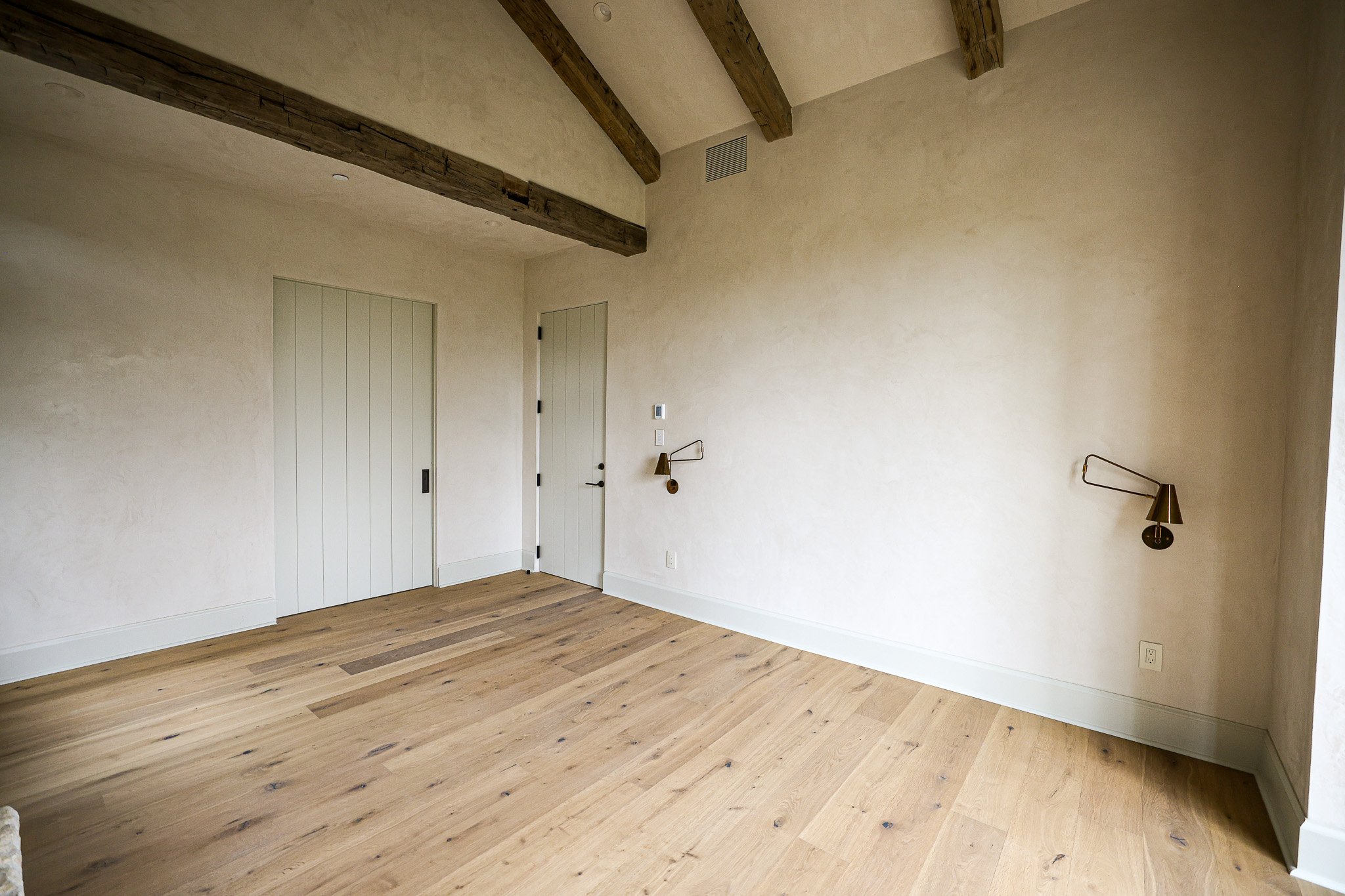 An empty room with cream-colored walls, wooden floor, exposed wooden ceiling beams, two white doors, wall-mounted reading lights, and a wall vent.