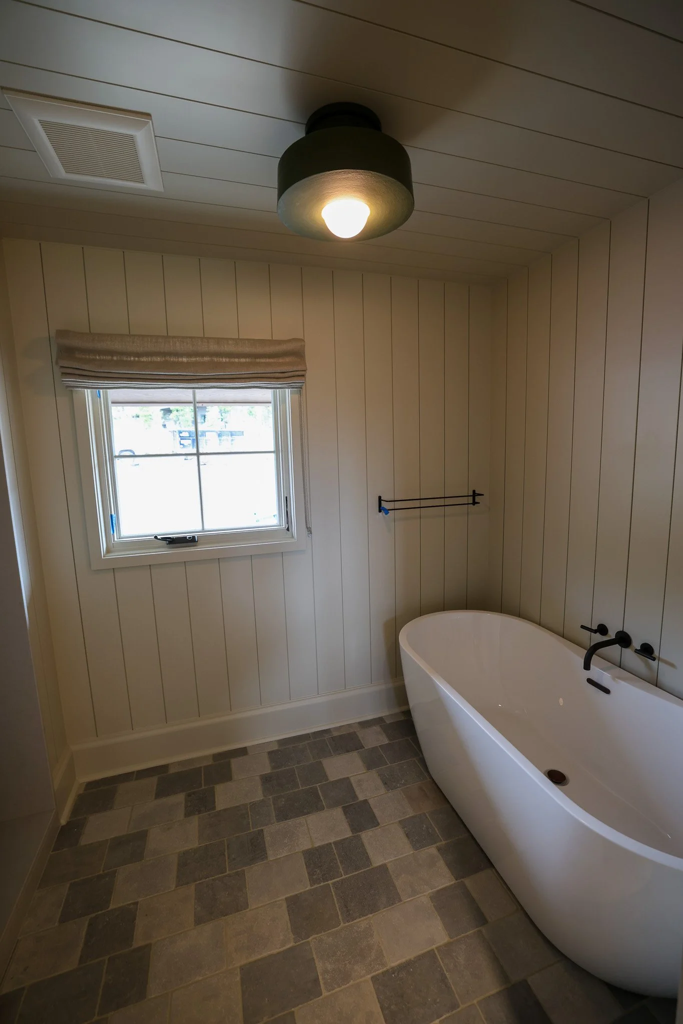 A bathroom with vertical wood paneling on the walls, a window with a beige Roman shade, a black towel rack, a black faucet, a white freestanding bathtub, a tile floor with various shades of gray, and a ceiling light fixture.