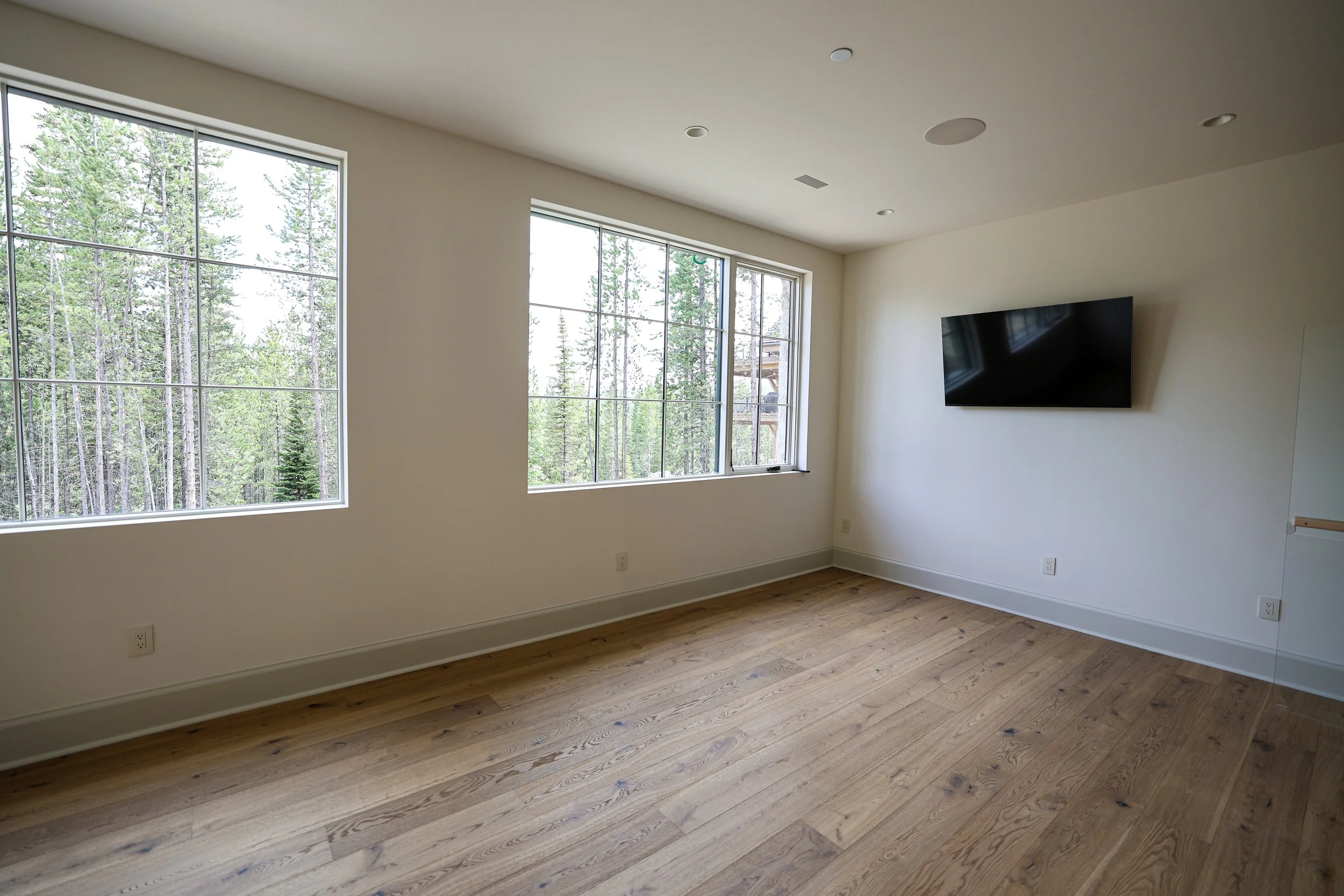 Empty room with two large windows showing a forest outside, mounted flat-screen TV on the wall, and hardwood floor.