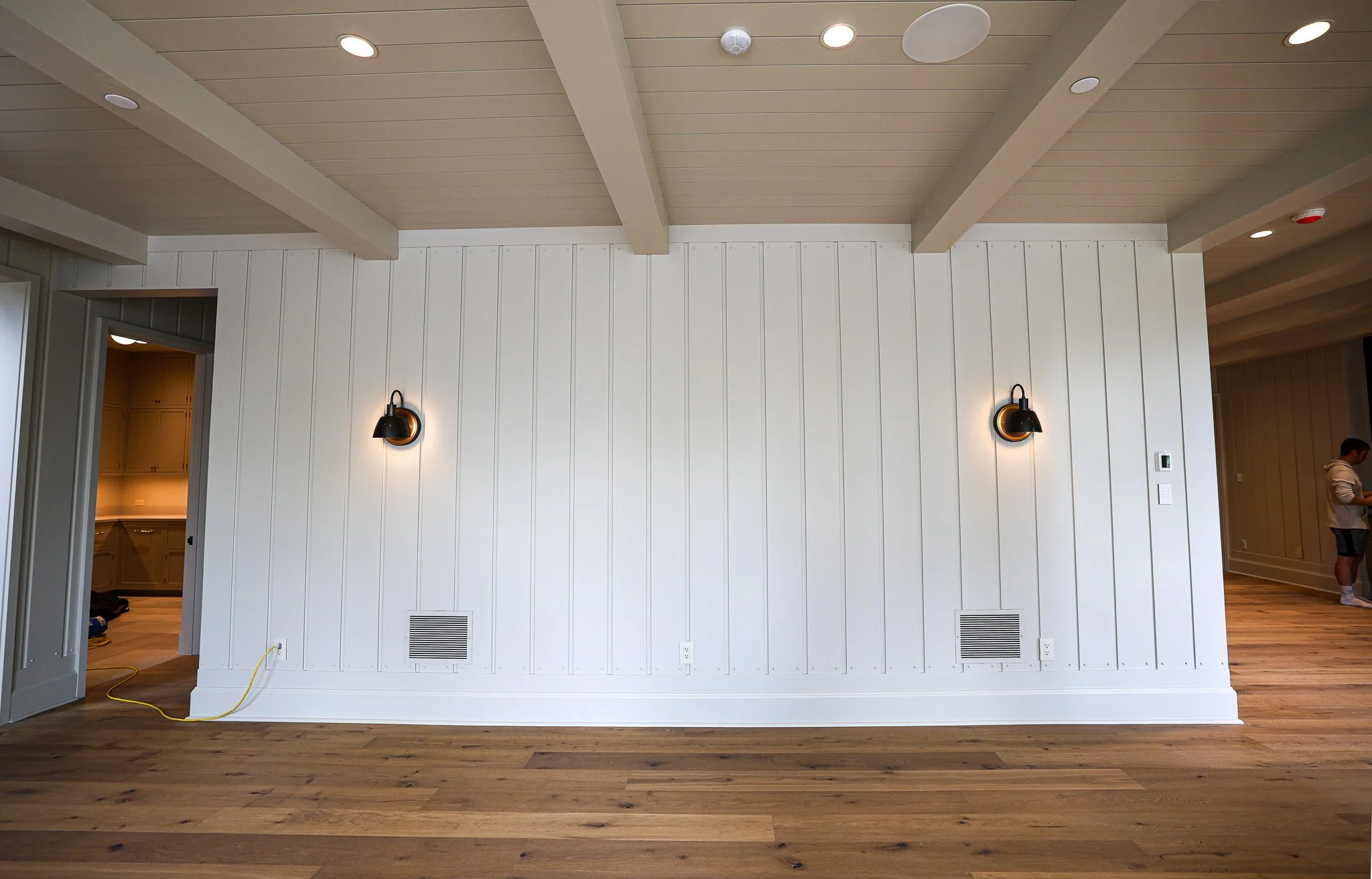 Interior view of a room with light-colored wood flooring and white walls with vertical paneling. Recessed ceiling lights and wall-mounted black and brass sconces are visible. A doorway leads to a kitchen area with yellow cabinetry. A person is partia