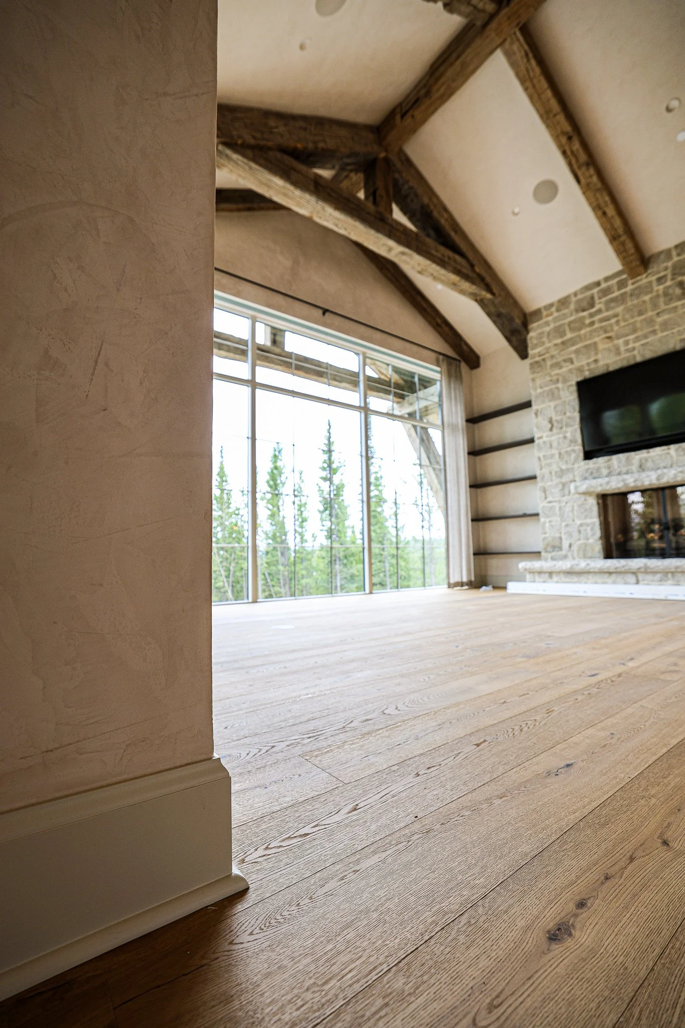 Interior of a room with wooden flooring, a stone fireplace, and large windows showing an outdoor view of trees. The ceiling has exposed wooden beams.
