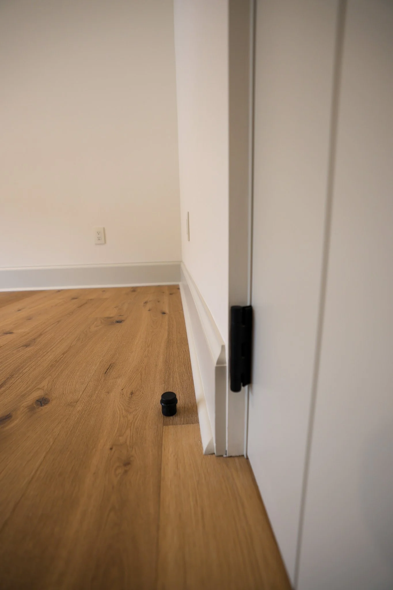 A corner of a room with hardwood flooring, a white wall with electrical outlets, and a white door with a black handle leaning or partially open. Two small black objects are on the floor near the door.