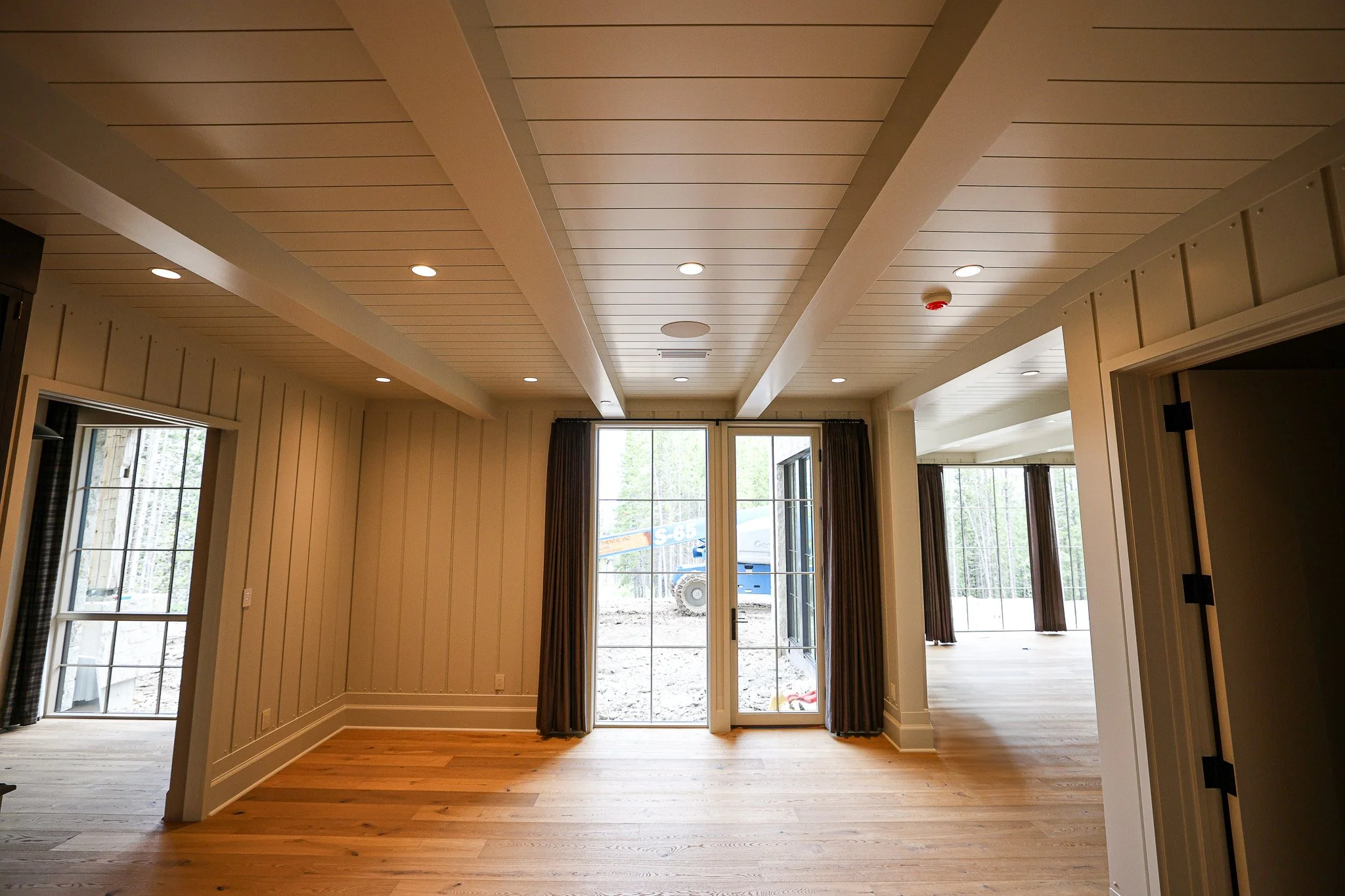 Interior of a house with cream-colored walls, hardwood floors, large glass sliding doors, and curtains, looking towards an outdoor area under construction.