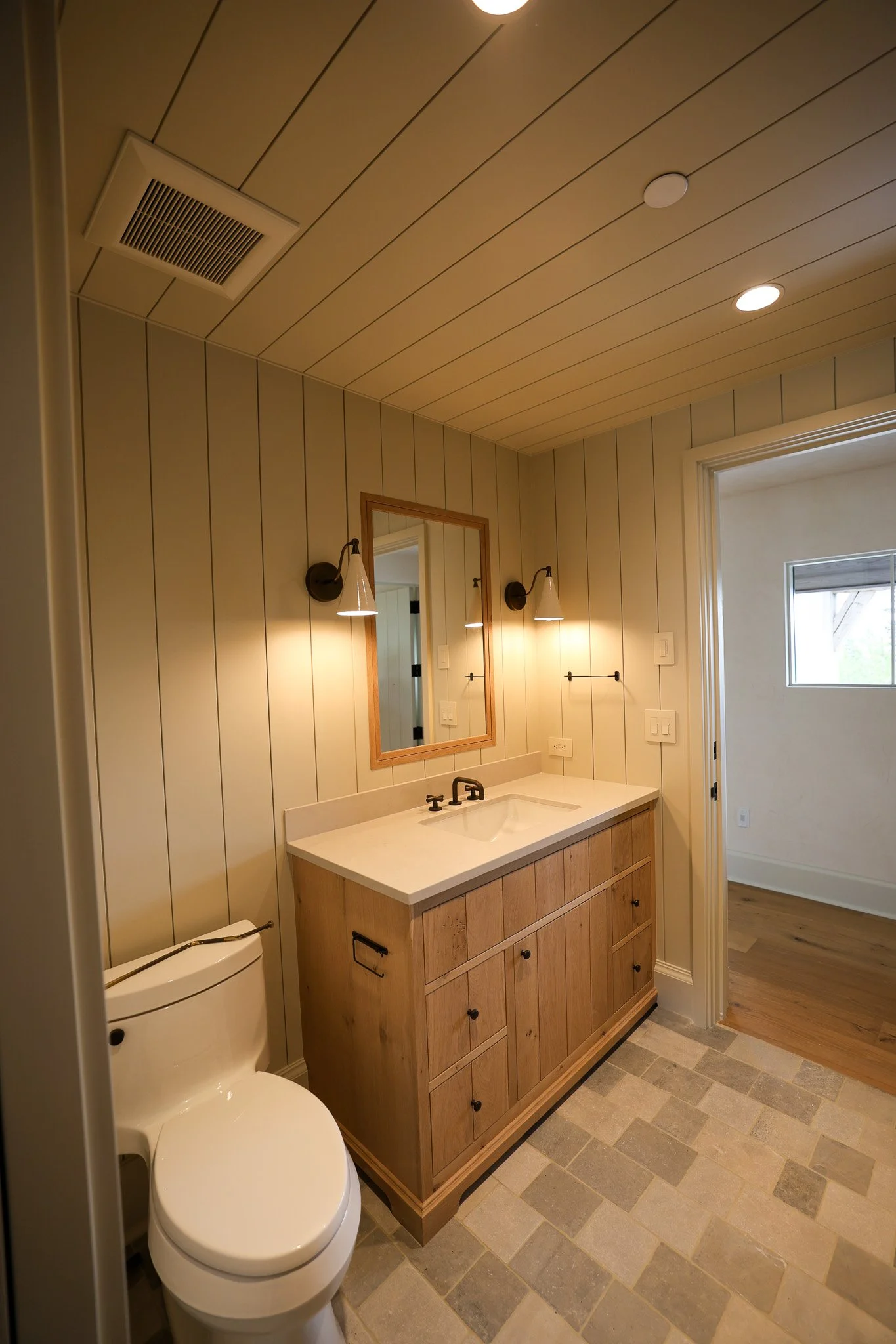 A bathroom with a wooden vanity, white countertop, and a rectangular mirror above. The wall is paneled with vertical wooden planks painted white. There is a toilet on the left, two wall-mounted light fixtures, and ceiling lights. The floor has beige and gray tiles, and there is an open doorway leading to a room with wooden flooring and a window.