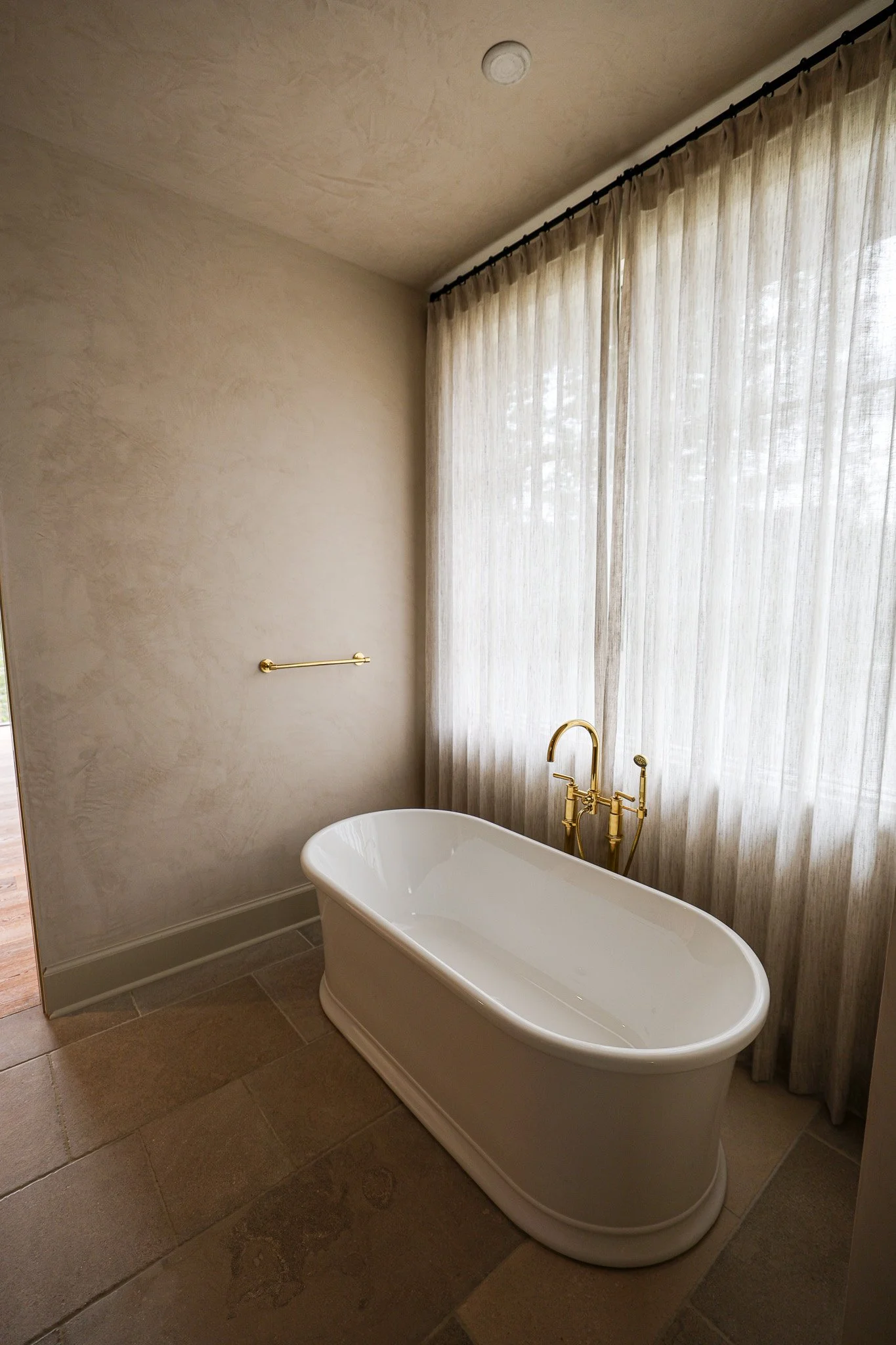 A white bathtub with gold fixtures in a bathroom, next to a window with sheer beige curtains, beige textured walls, and light brown tiled floor.