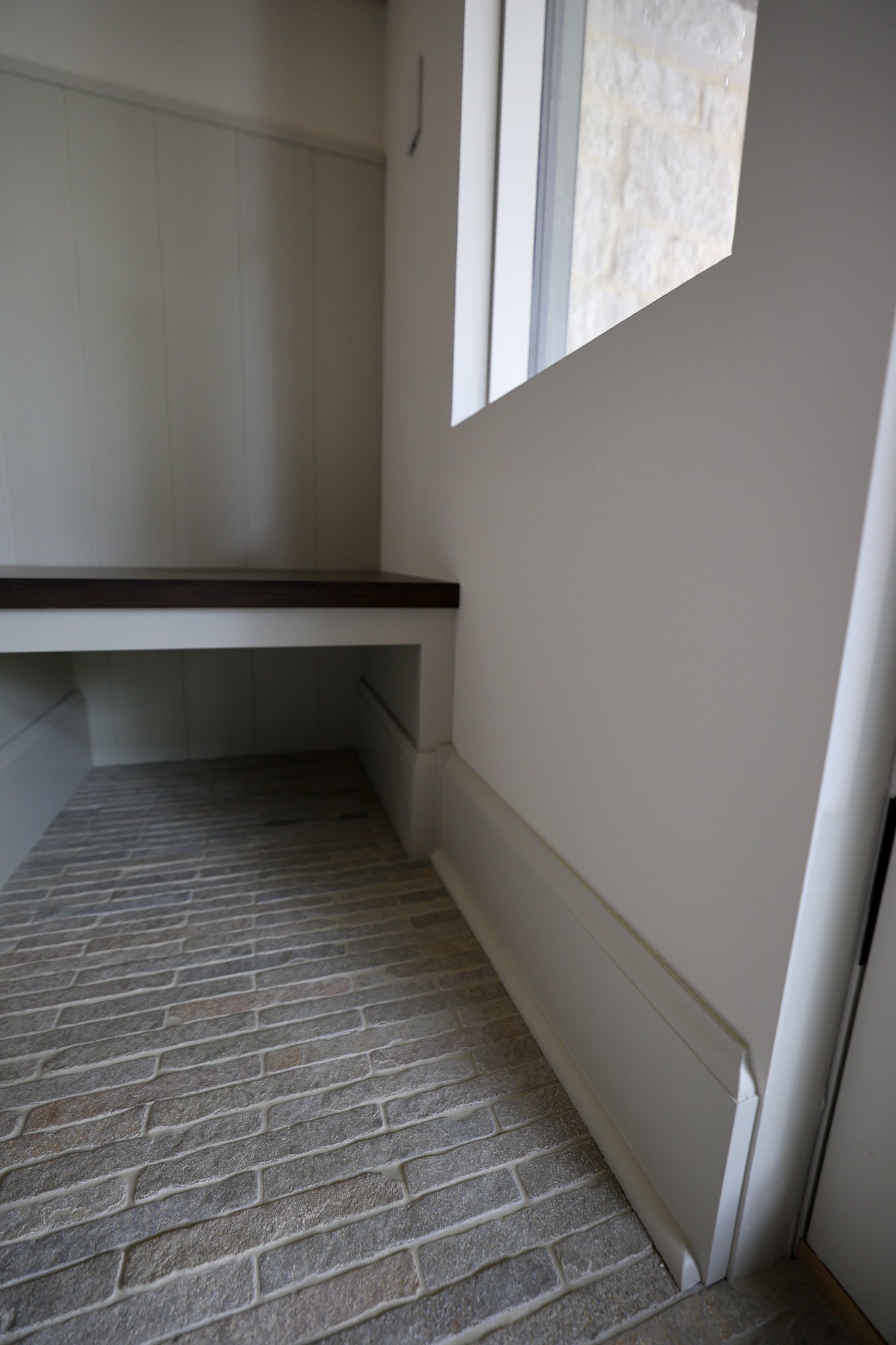 Interior view of a room with beige walls, a window with stone wall visible outside, a dark wooden ledge, and a brick-like textured floor.