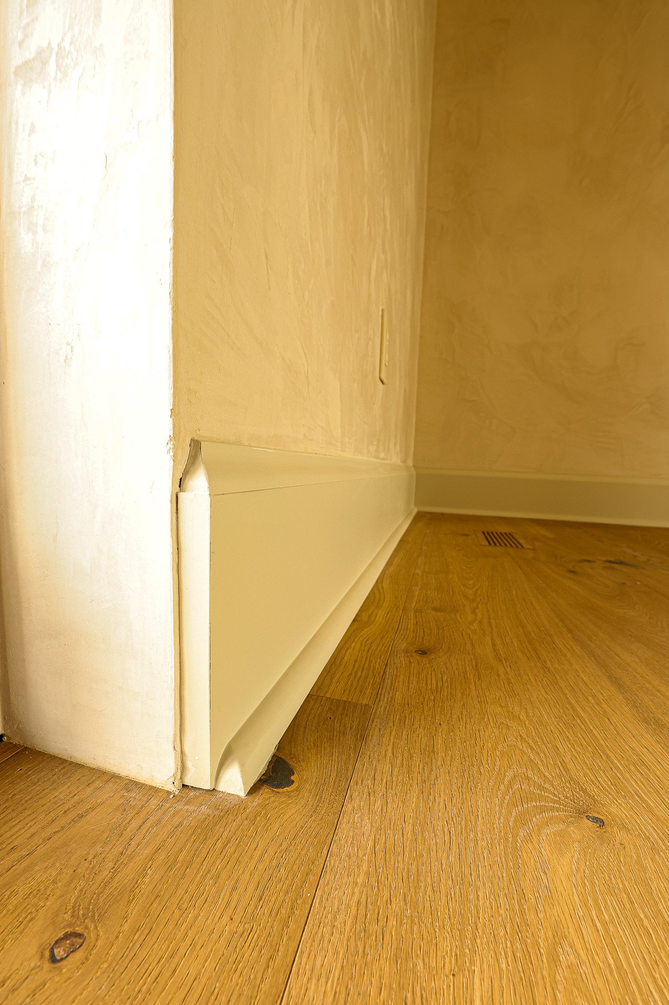 Close-up of a corner where a white baseboard meets a beige wall and wooden floor.
