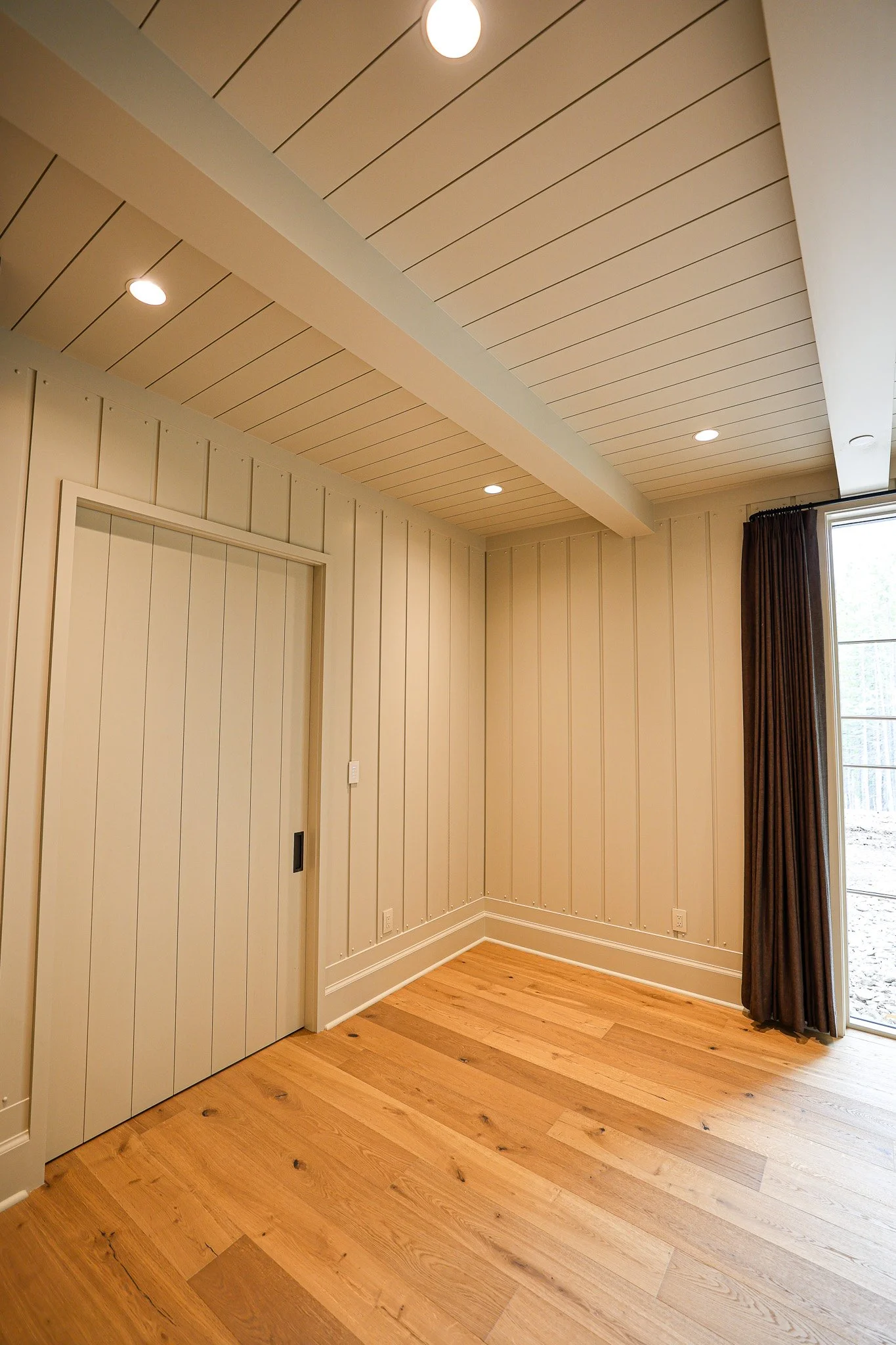 A corner of a room featuring light beige wood paneling on the walls and ceiling, with a door flush to the wall, a window with dark curtains, and a hardwood floor.