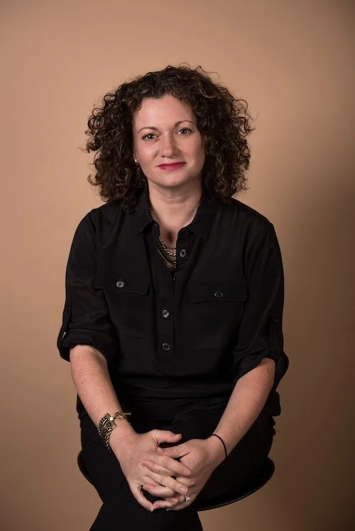 Image of Judith Pineiro, a museum and global art-world consultant.  Sitting on a stool against a beige background, wearing a black button-up shirt and gold jewelry.