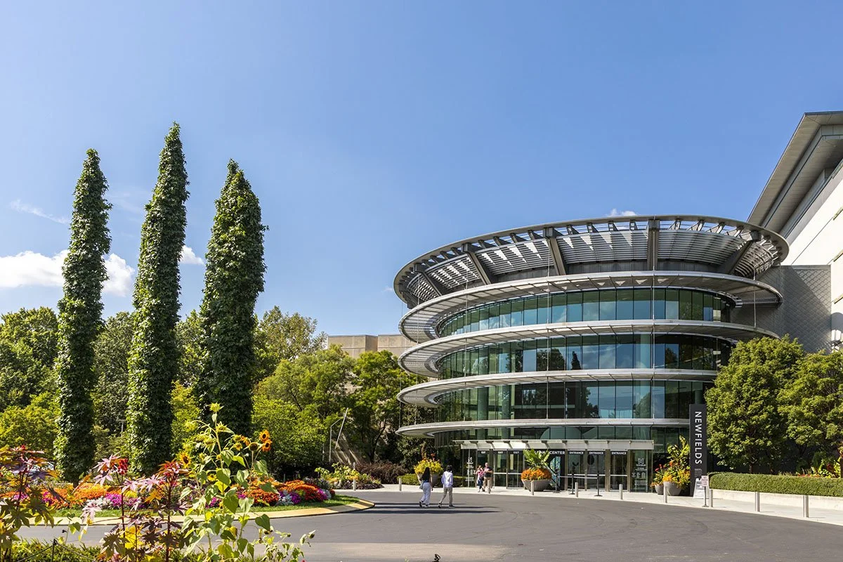 Exterior view of the Newfields art and gardens center with modern glass and metal architecture, surrounded by trees and colorful flowers on a sunny day.