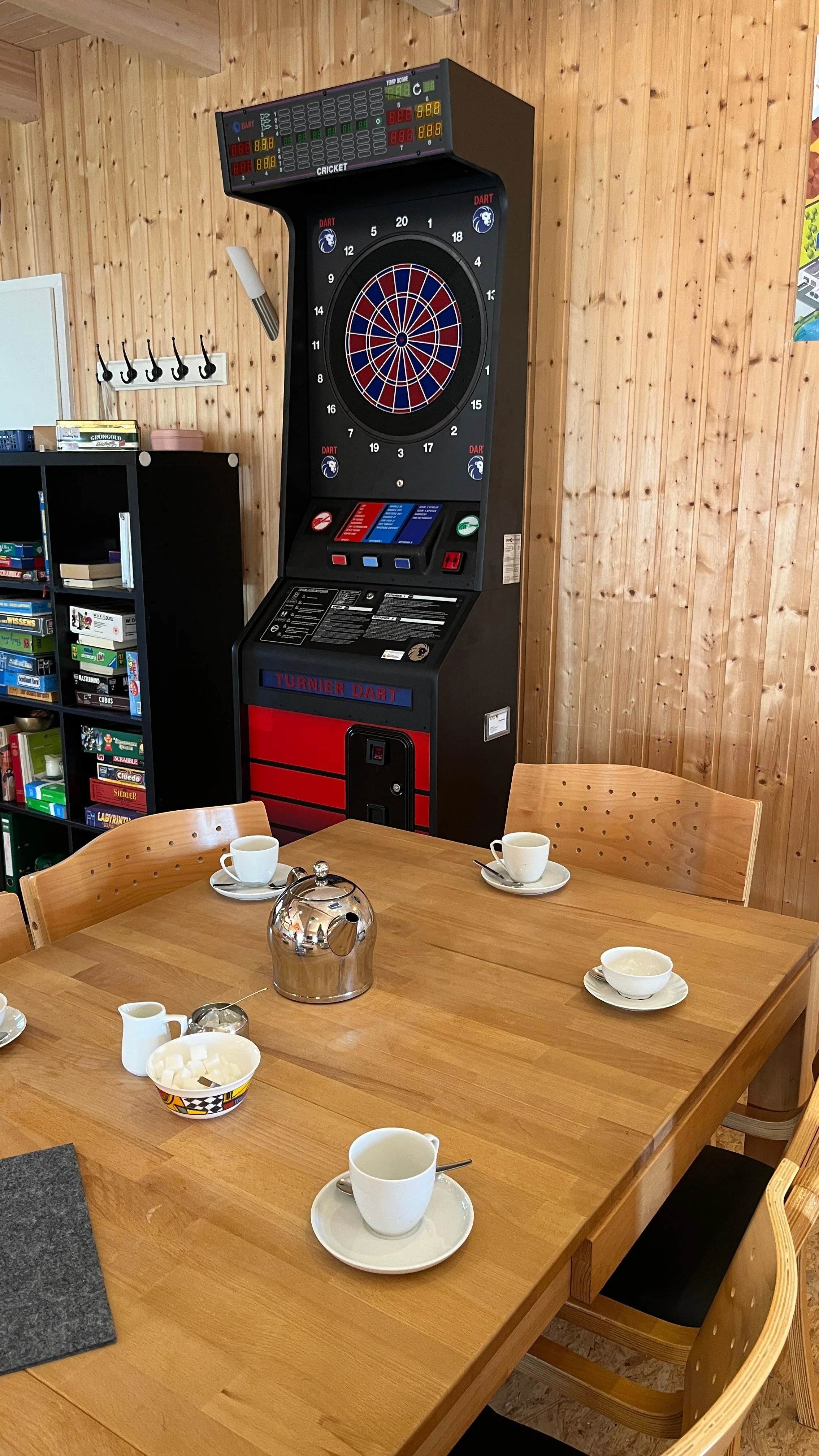 Ein Dartautomat in einem Raum mit Holzvertäfelung, umgeben von einem Holztisch mit Kaffeetassen, einem Teekessel und Zucker.
