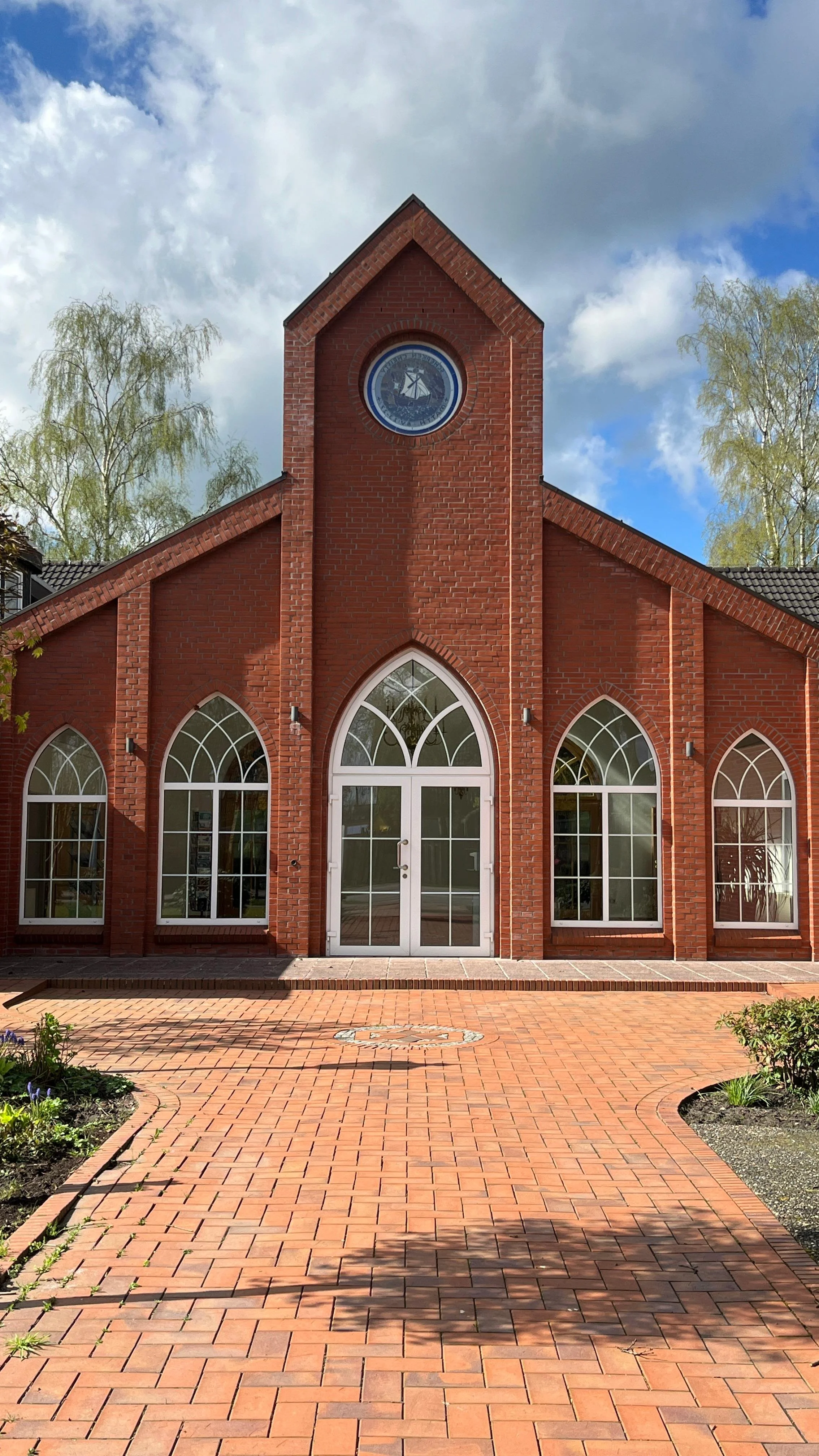 Rotes Backsteingebäude mit großen Fensterbögen und einer Uhr über dem Eingang, umgeben von einem gepflasterten Vorplatz und Bäumen unter einem bewölkten Himmel.