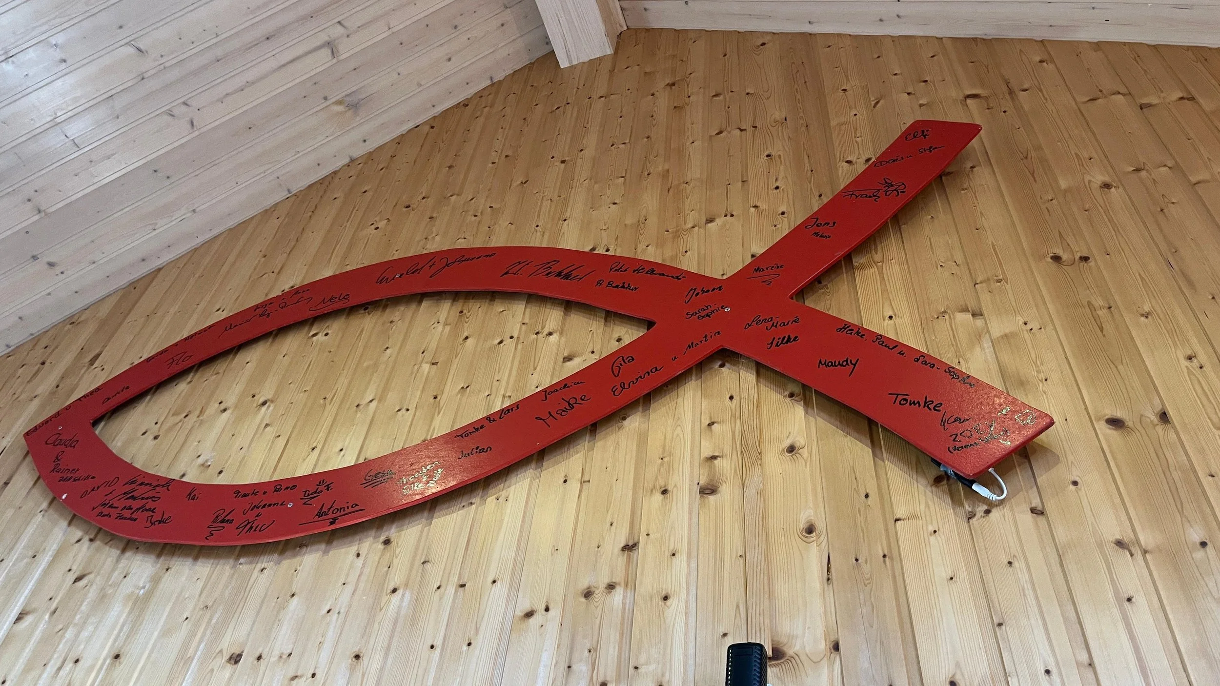Ein großes, rot bemaltes Holzschild in Form einer Fischform, an der zahlreiche Unterschriften und Grüße in Schwarz geschrieben sind, an einer Holzvertäfelung im Raum befestigt.
