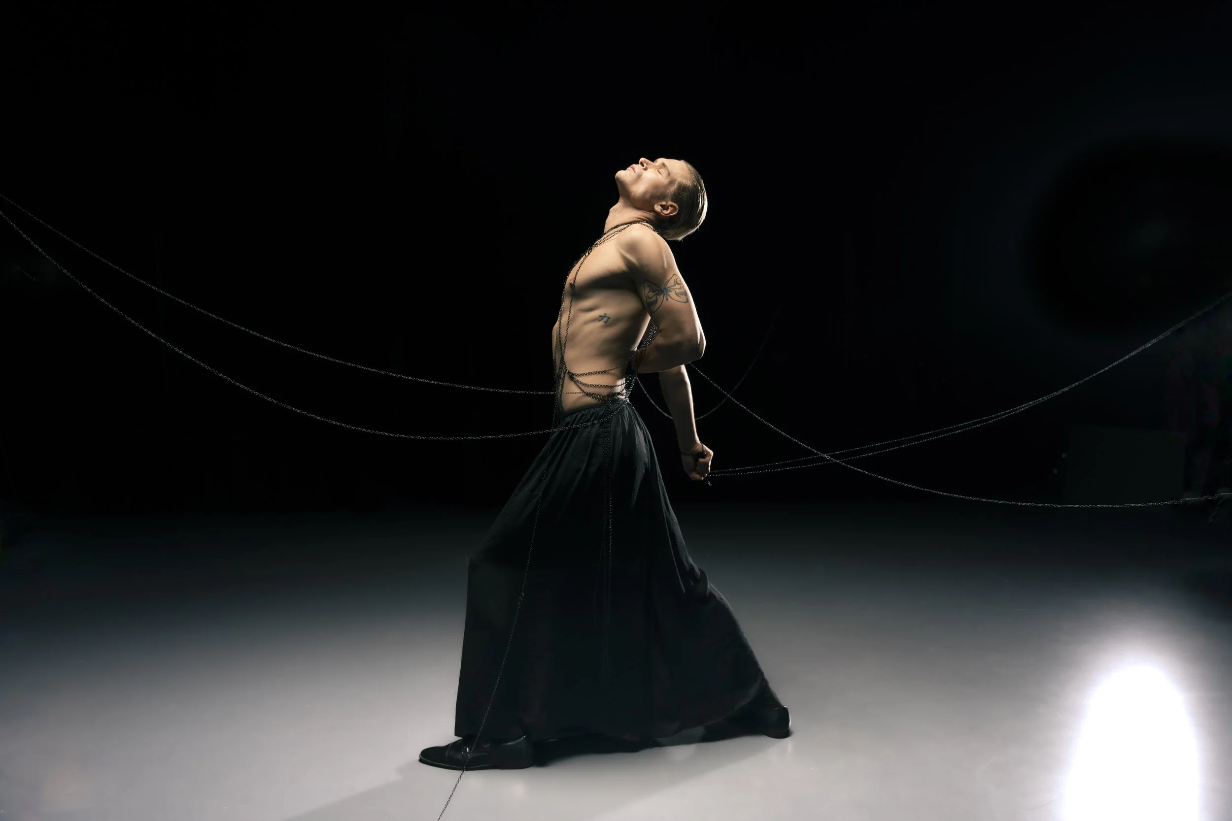 Ein männlicher Tänzer steht im dunklen Raum, mit Blick nach oben, nackt bis auf eine schwarze, eleganten Hose, mit Tattoos an den Armen und einem langen schwarzen Schal oder Tuch, das um seinen Körper gewickelt ist, in einer dramatischen Pose.