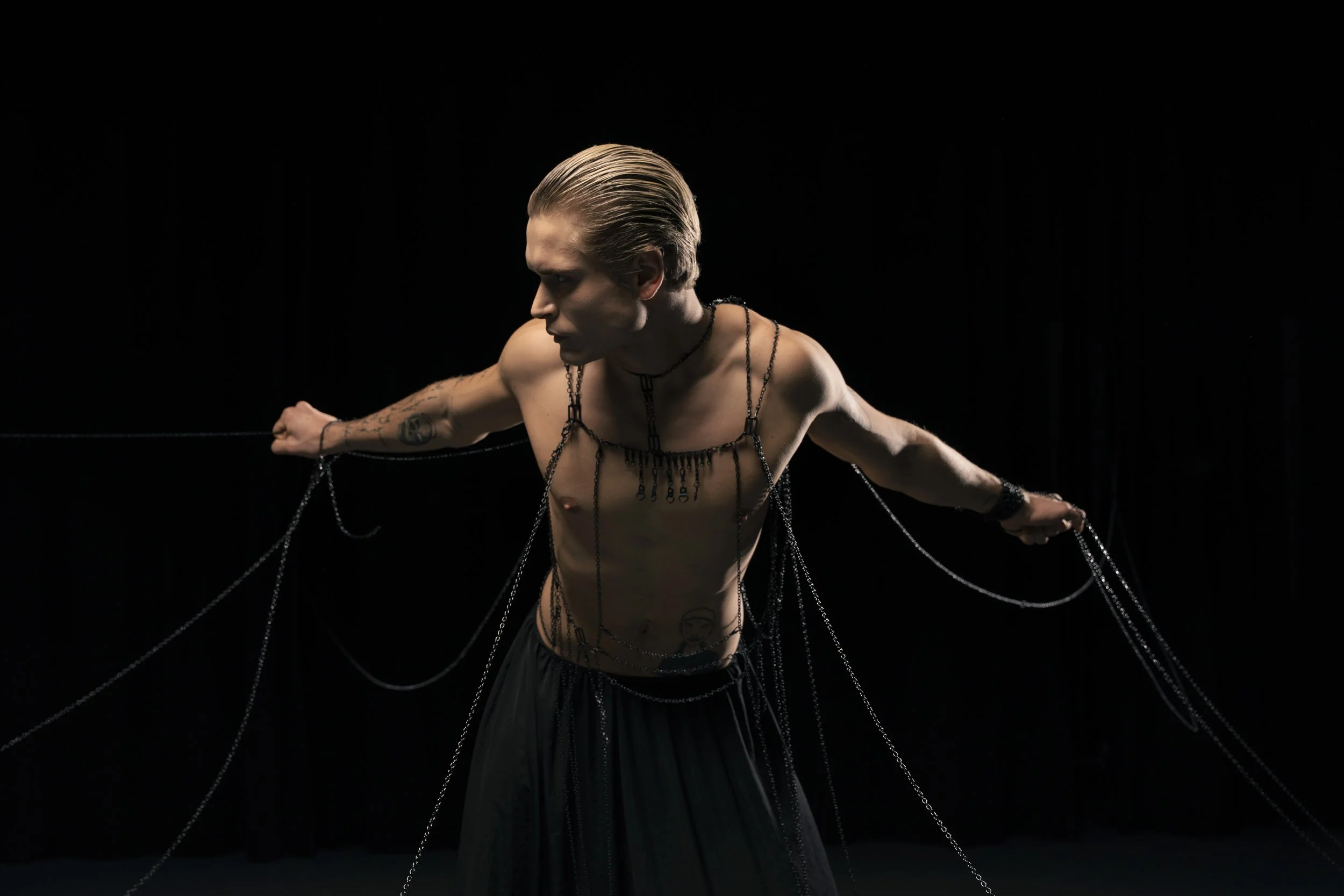 A shirtless man with slicked-back hair is holding chains that are attached to his arms, set against a black background.