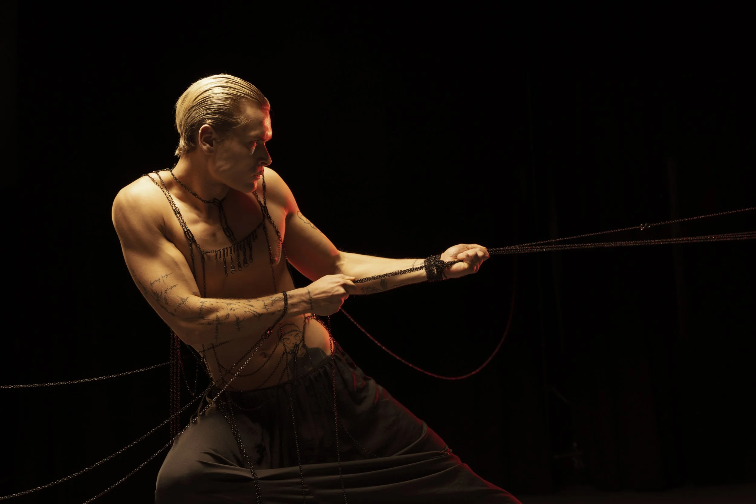 A shirtless man with tattoos and light blond hair, illuminated by red and yellow lighting, holding multiple chains or ropes against a black background.