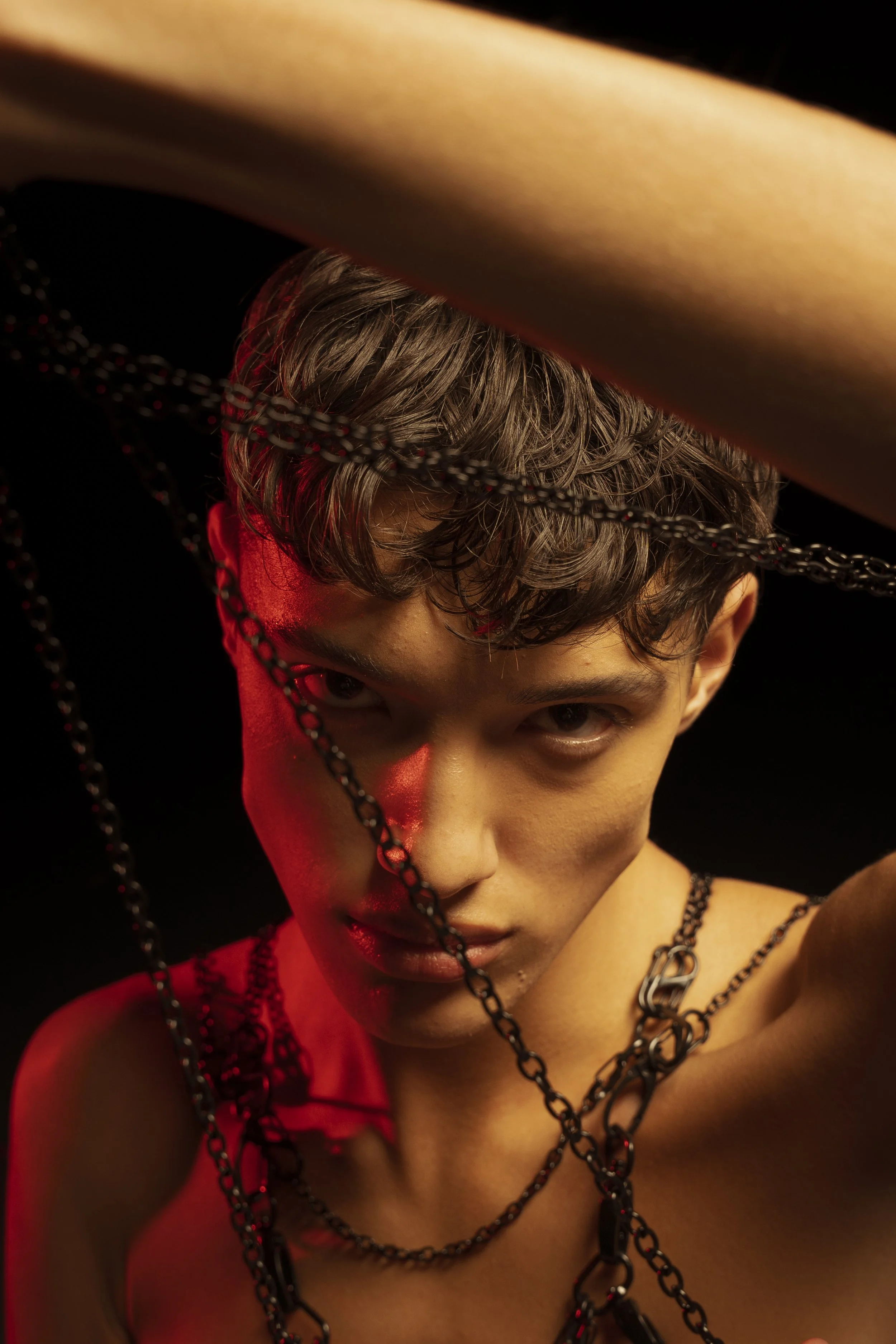 Junge Person mit dunklen Haaren und Ketten vor schwarzem Hintergrund, teilweise von oben betrachtet, mit rotem Licht betont.