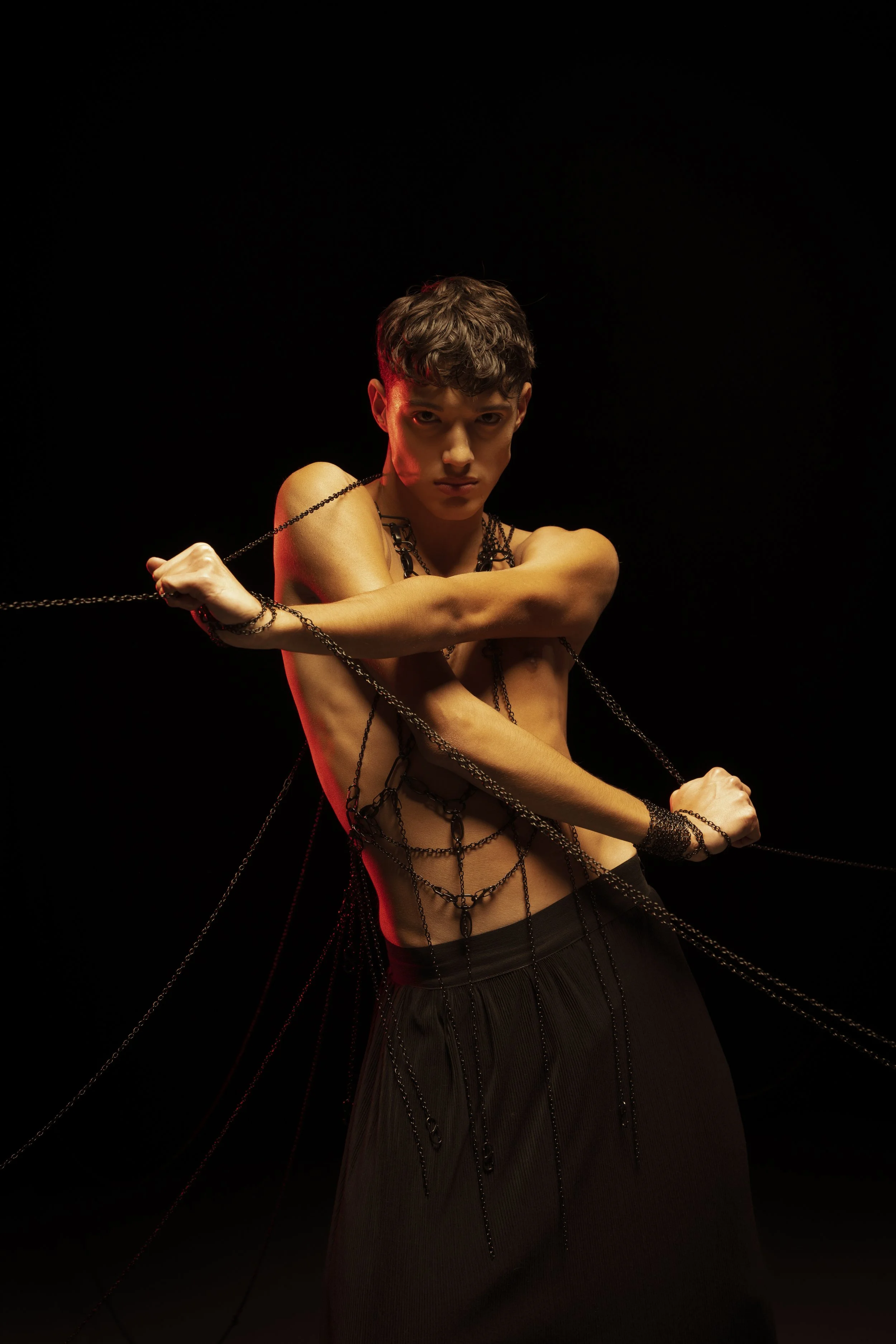 Junger Mann in dunkler Kleidung und mit schwarzen Ketten bedeckt, posiert dramatisch vor einem schwarzen Hintergrund.