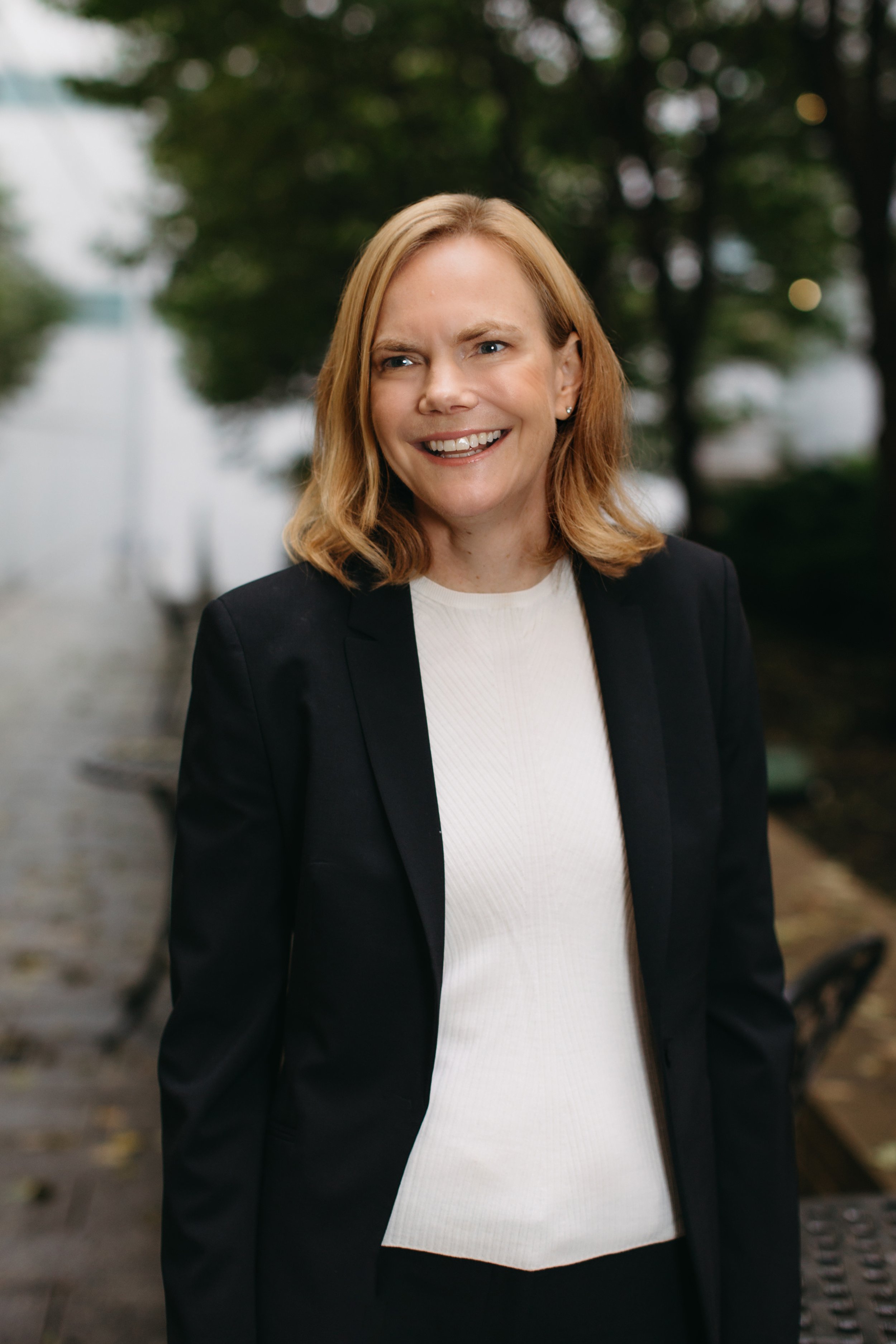 A woman with red hair, wearing a black blazer and white top, smiling outdoors with trees in the background.