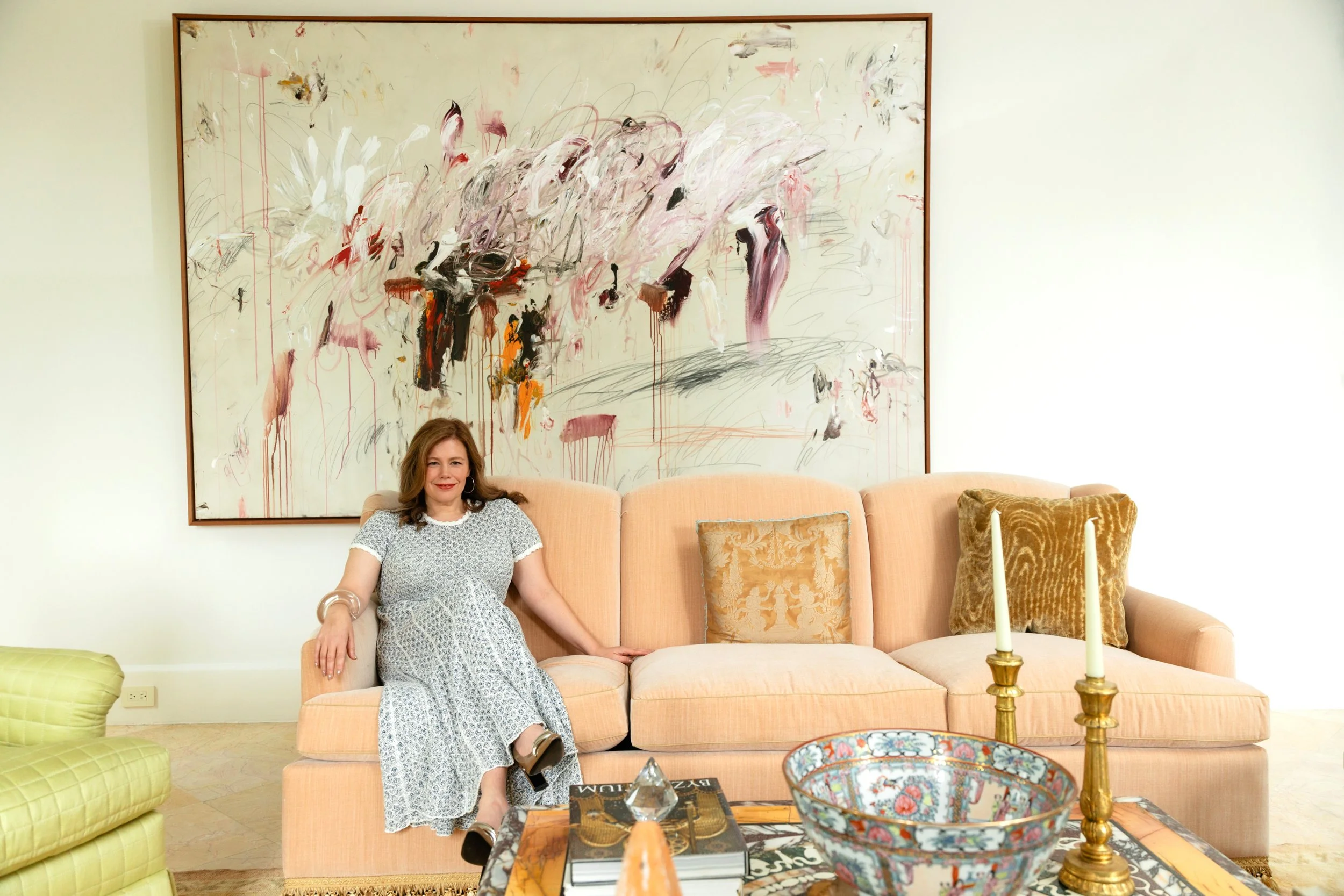 A woman with long brown hair sitting on a peach-colored sofa in a living room, with colorful abstract art hanging on the wall behind her. She is smiling and wearing a white patterned dress. There are decorative pillows on the sofa, a coffee table with books and a decorative bowl, and gold candlesticks with candles on a side table.