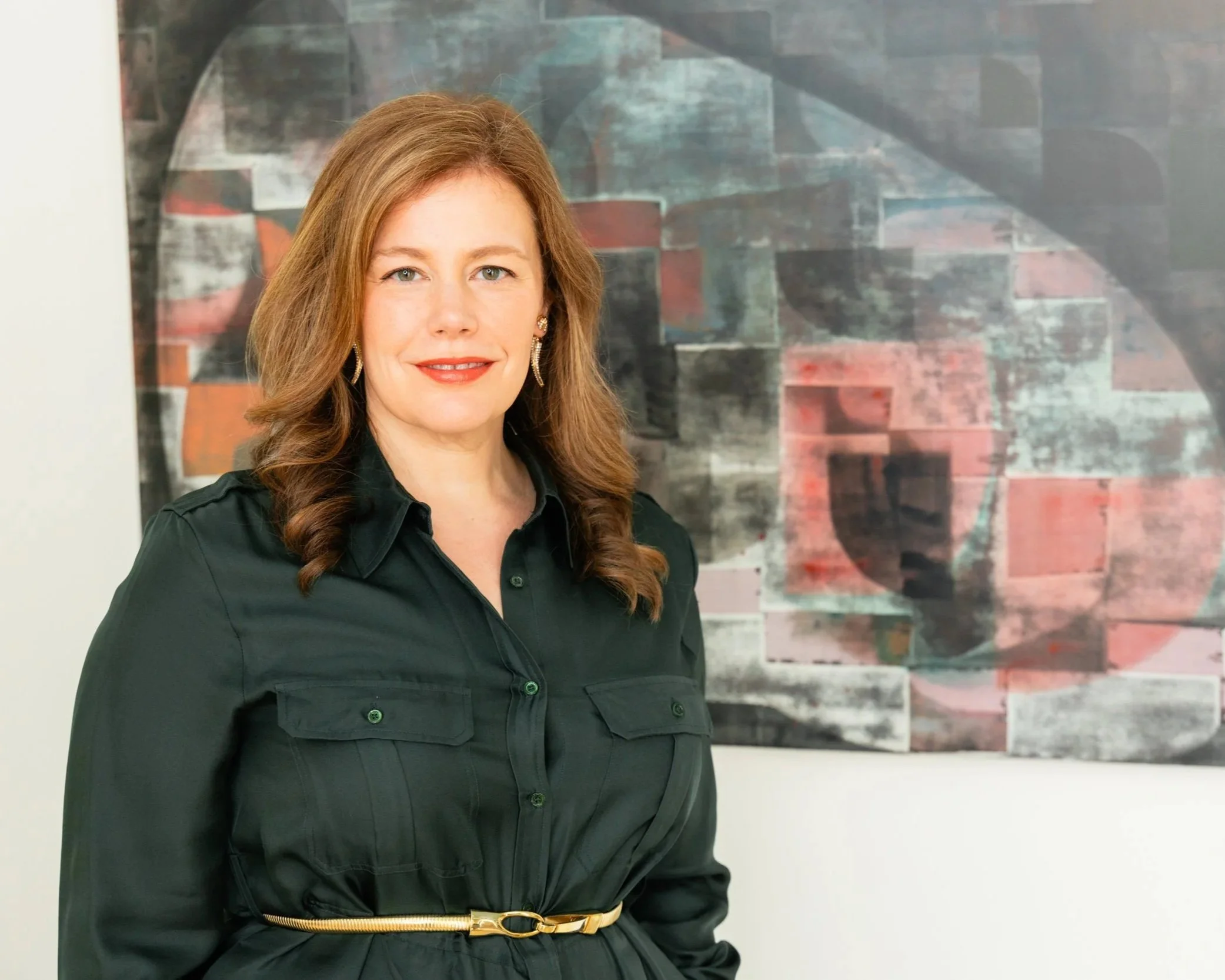 A woman with long, wavy, light brown hair and blue eyes, smiling, wearing a black shirt and a belt, standing in front of or near a colorful abstract artwork.