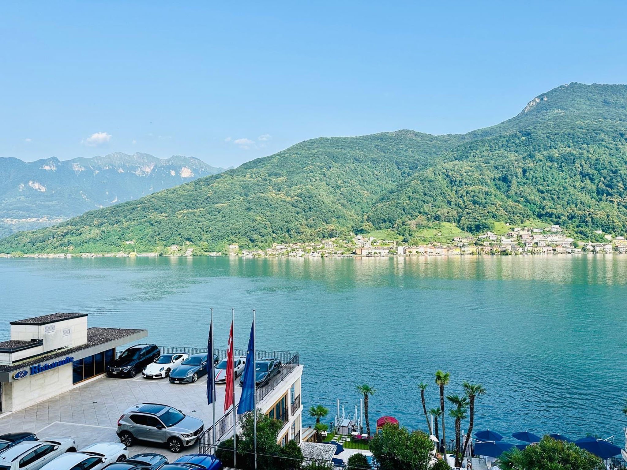 See mit umliegenden grünen Bergen im Sonnenschein, Gebäude und Autos im Vordergrund, Palmen und Sonnenschirme entlang des Wassers, klarer blauer Himmel im Hintergrund.