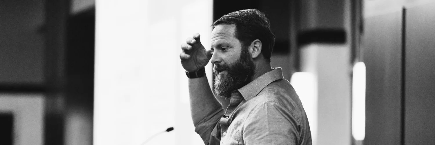 A man with a beard and short hair, wearing a button-up shirt, appears to be giving a presentation or speech, standing in a room with a microphone and a blurred background.