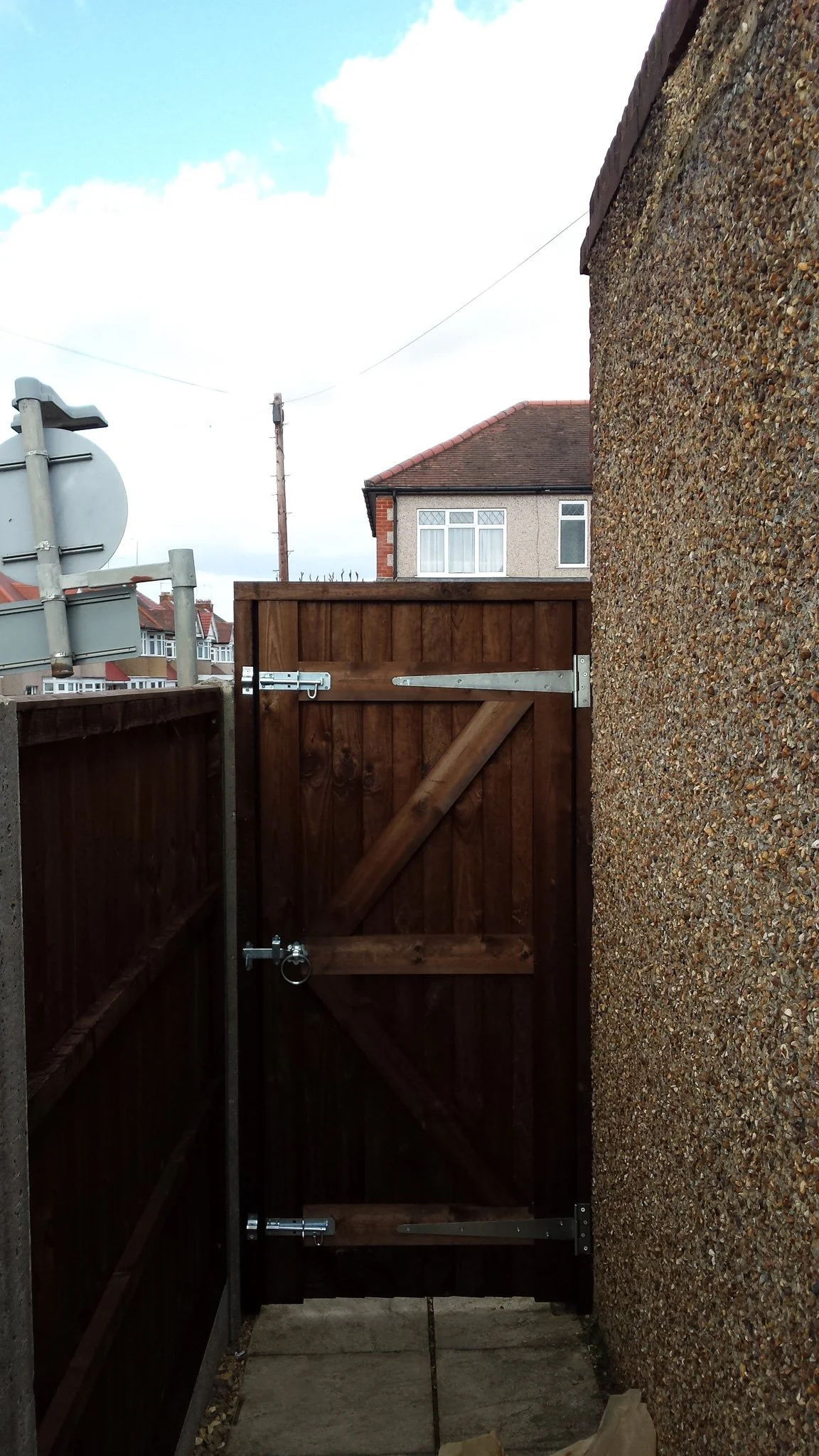 A wooden garden gate with metal hinges and a latch, between a pebble-dash wall and a wooden fence, with a residential street and houses visible in the background.
