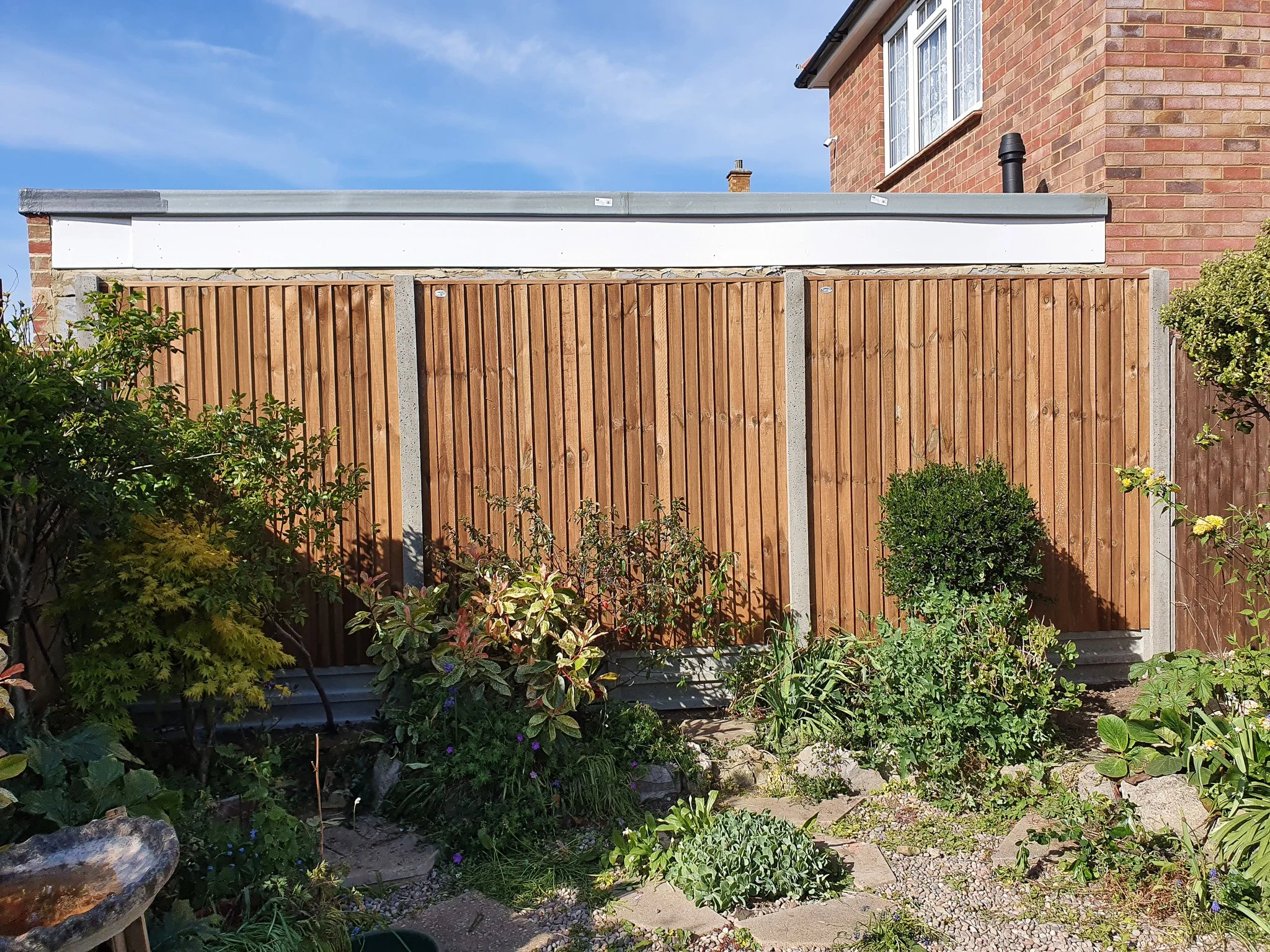 Wooden garden fence with plants and bushes in front of it, part of a brick house visible on the right, blue sky above.