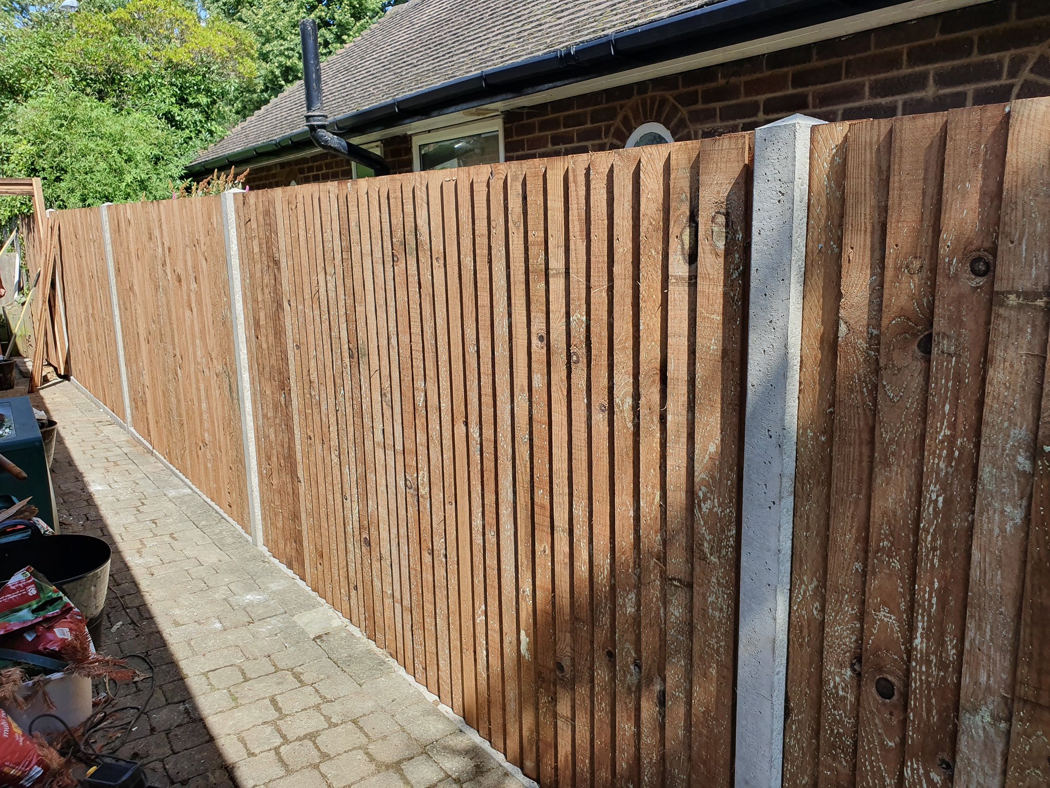 A newly installed wooden fence along a brick walkway in a backyard, with a house and trees in the background.