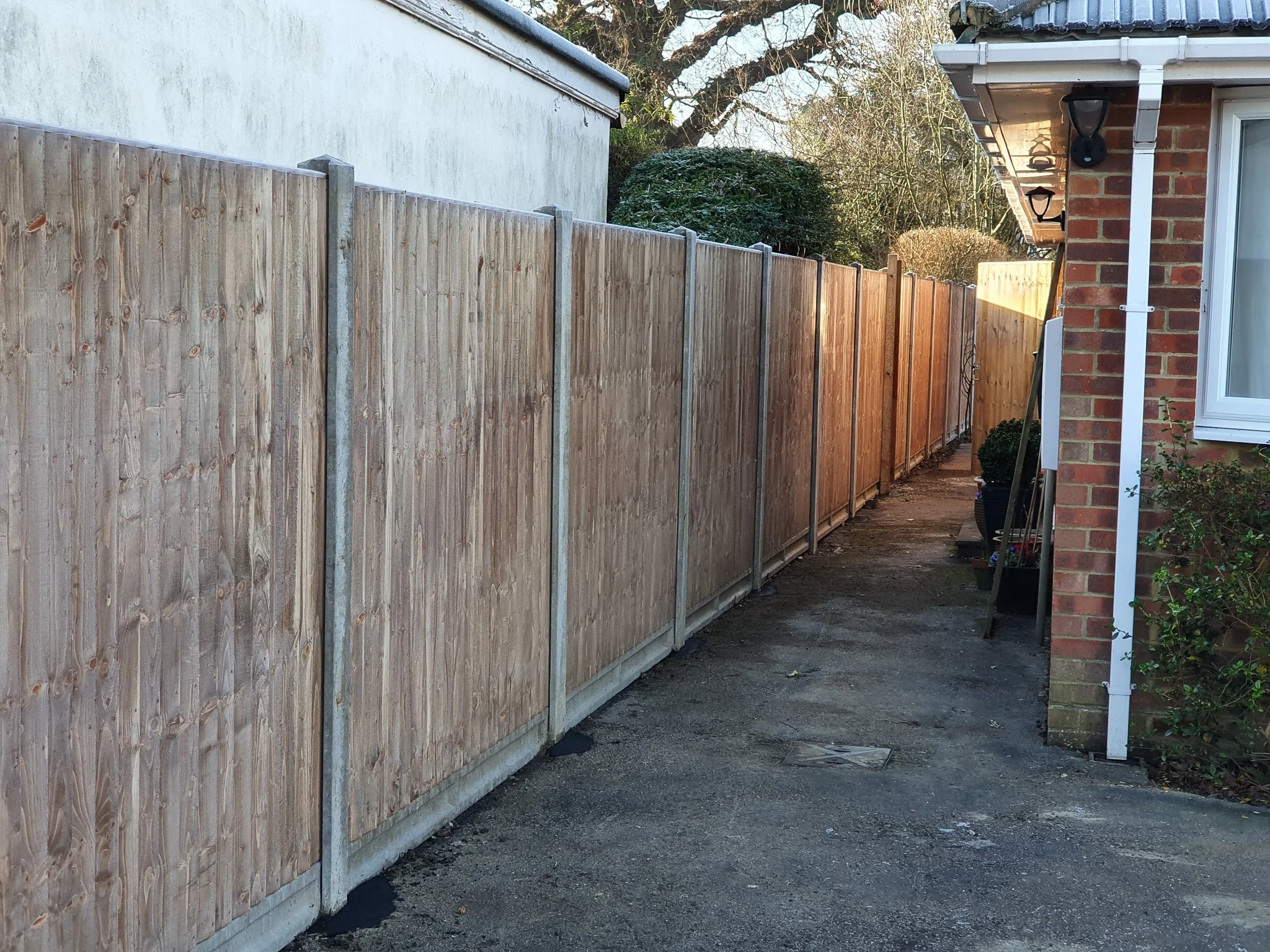 A tall wooden fence runs along the side of a house, with a paved pathway beside it. The house has a brick exterior with black outdoor wall lights and a white gutter. There are potted plants and gardening tools near the house.
