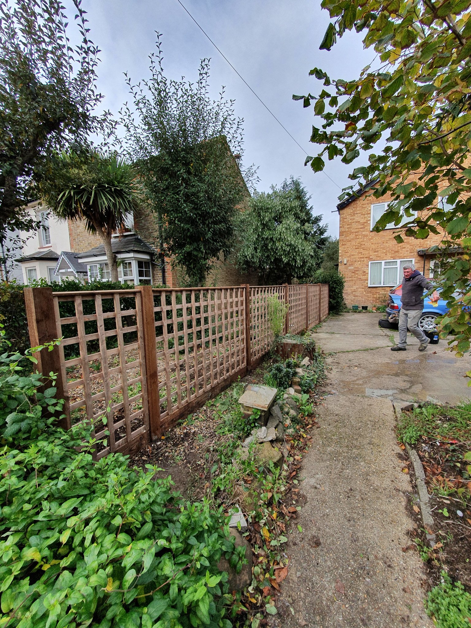 A man walking on a concrete driveway next to a freshly installed wooden fence, with a garden and trees on the left and a brick house on the right, under a cloudy sky.