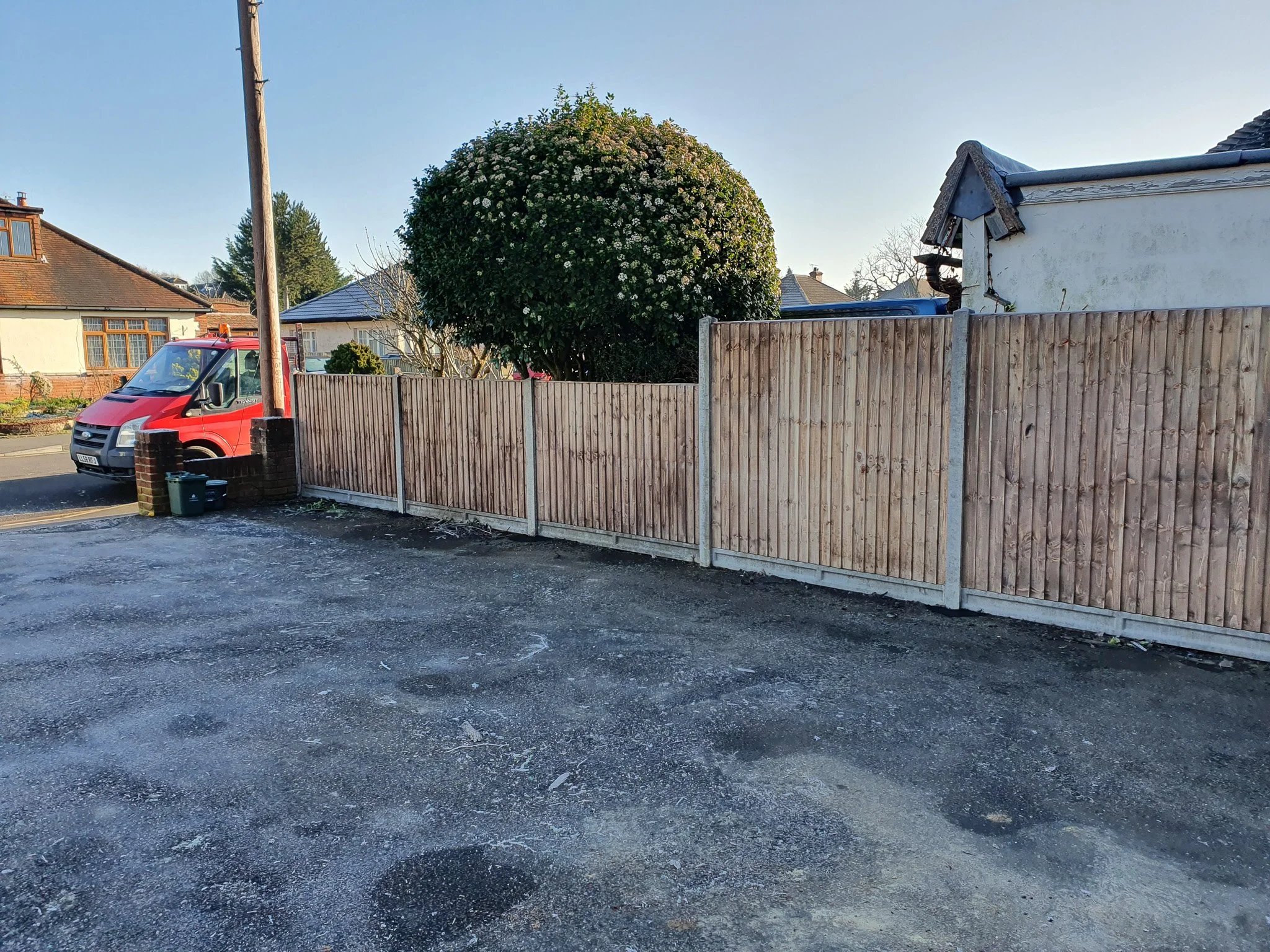 A residential street scene with a red van parked on the road, a wooden fence dividing properties, a large tree with white flowers, and houses in the background under clear blue sky.