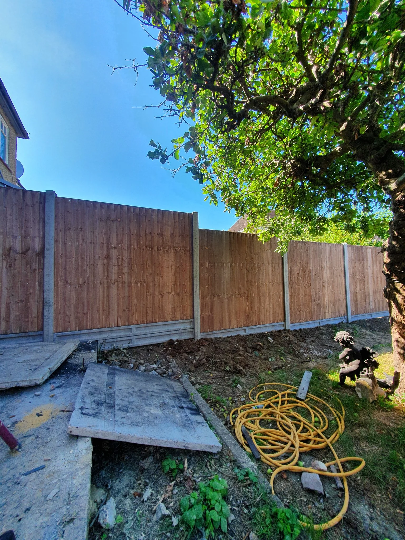 A backyard with a wooden fence, a tree with green leaves, a blue sky, construction materials, including a concrete slab, a yellow hose, and a statue of a gnome.