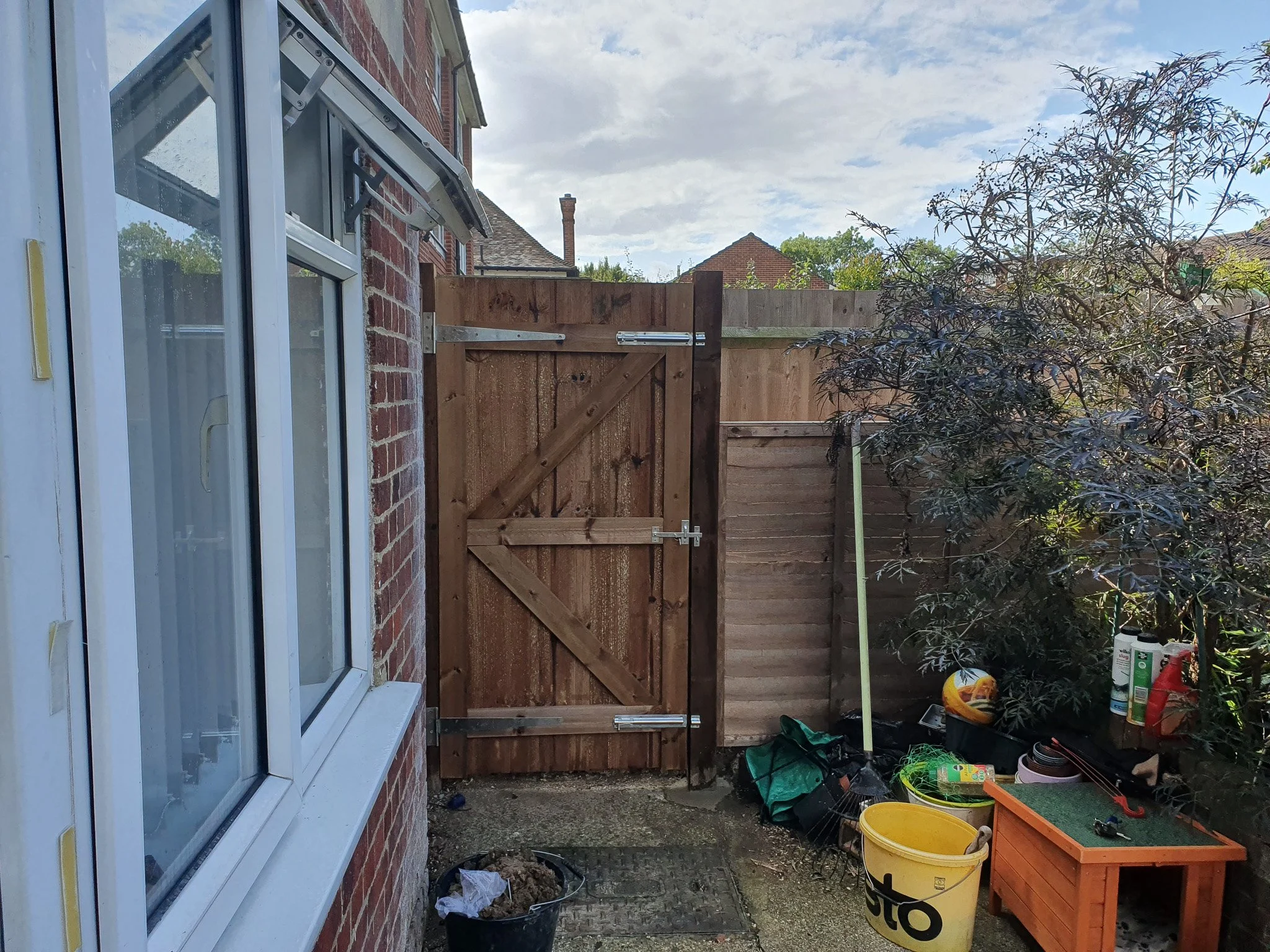 Small backyard with wooden fencing, a bush on the right, and gardening supplies including a yellow bucket, a small orange planter, and various tools.