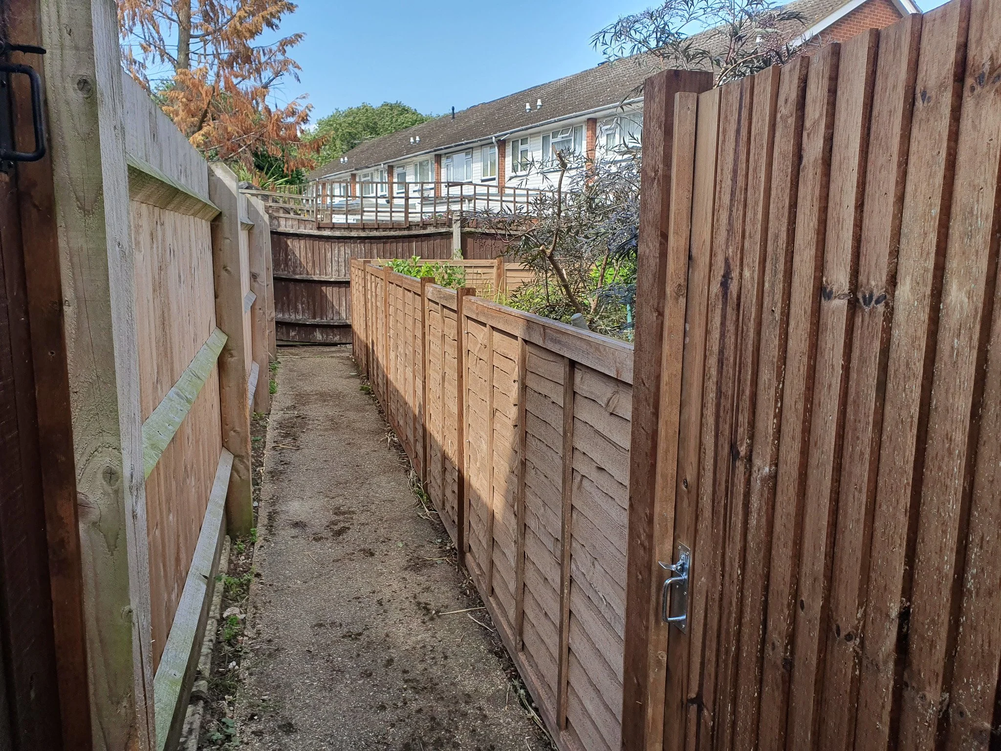 A narrow backyard pathway bordered by a newly installed wooden fence on the right and an existing wooden fence on the left. The pathway leads toward a gate at the back with houses visible beyond.