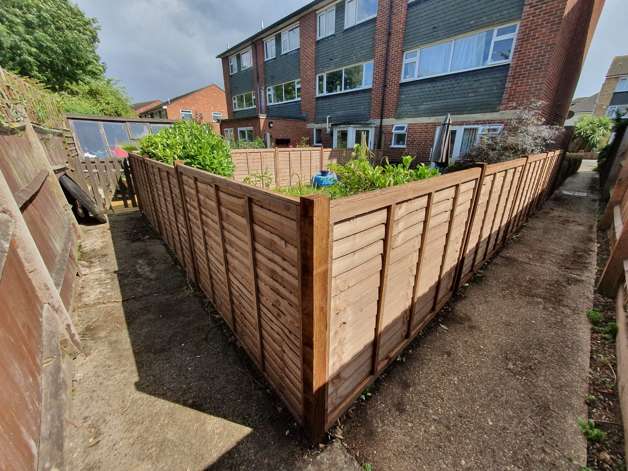 A backyard garden enclosed by wooden fencing, with plants and outdoor furniture, in a residential area with brick apartment buildings.