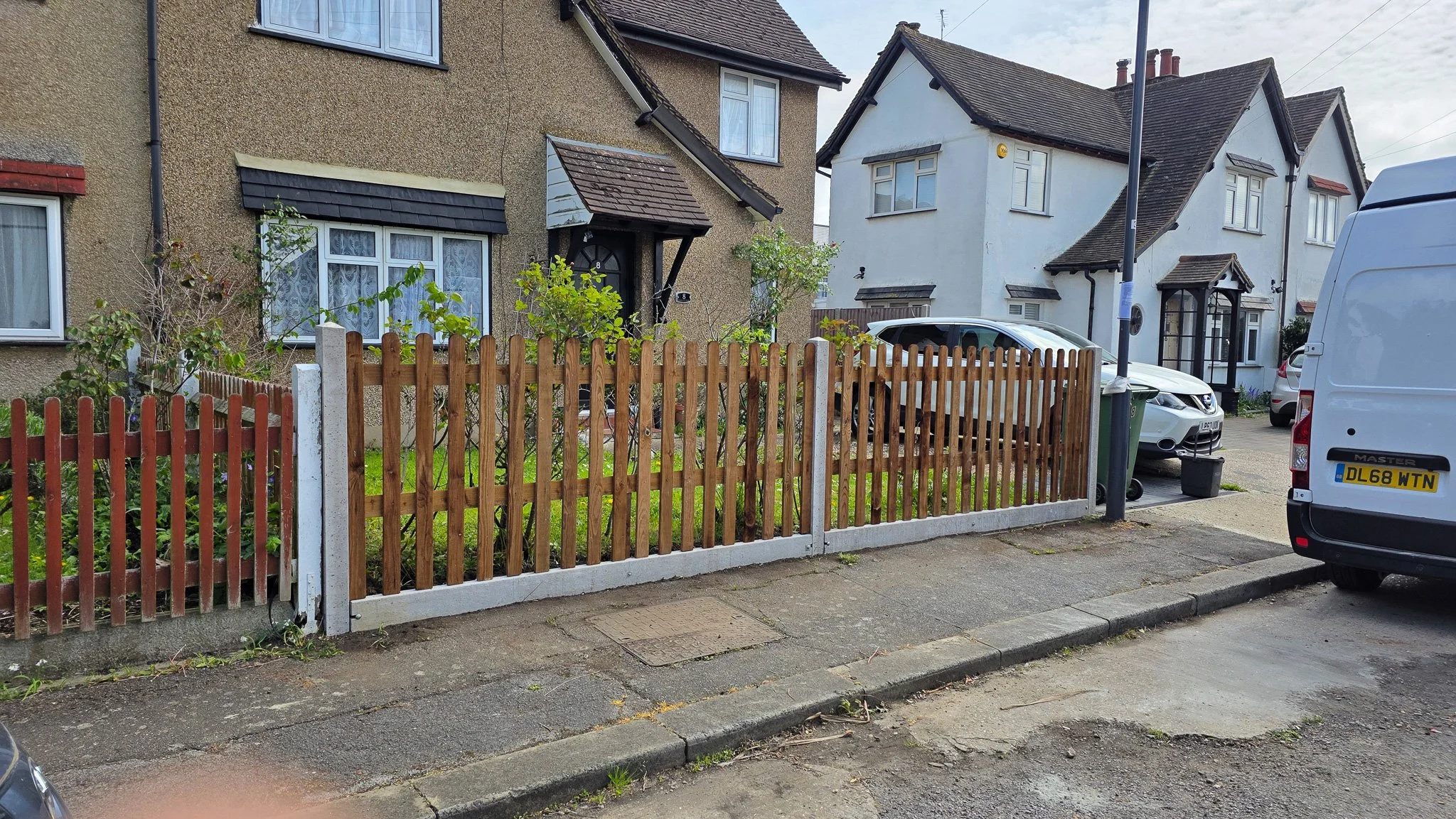 Residential street scene with a house having a garden and a wooden fence in front, parked cars, trash bins, a streetlight, and neighboring houses.