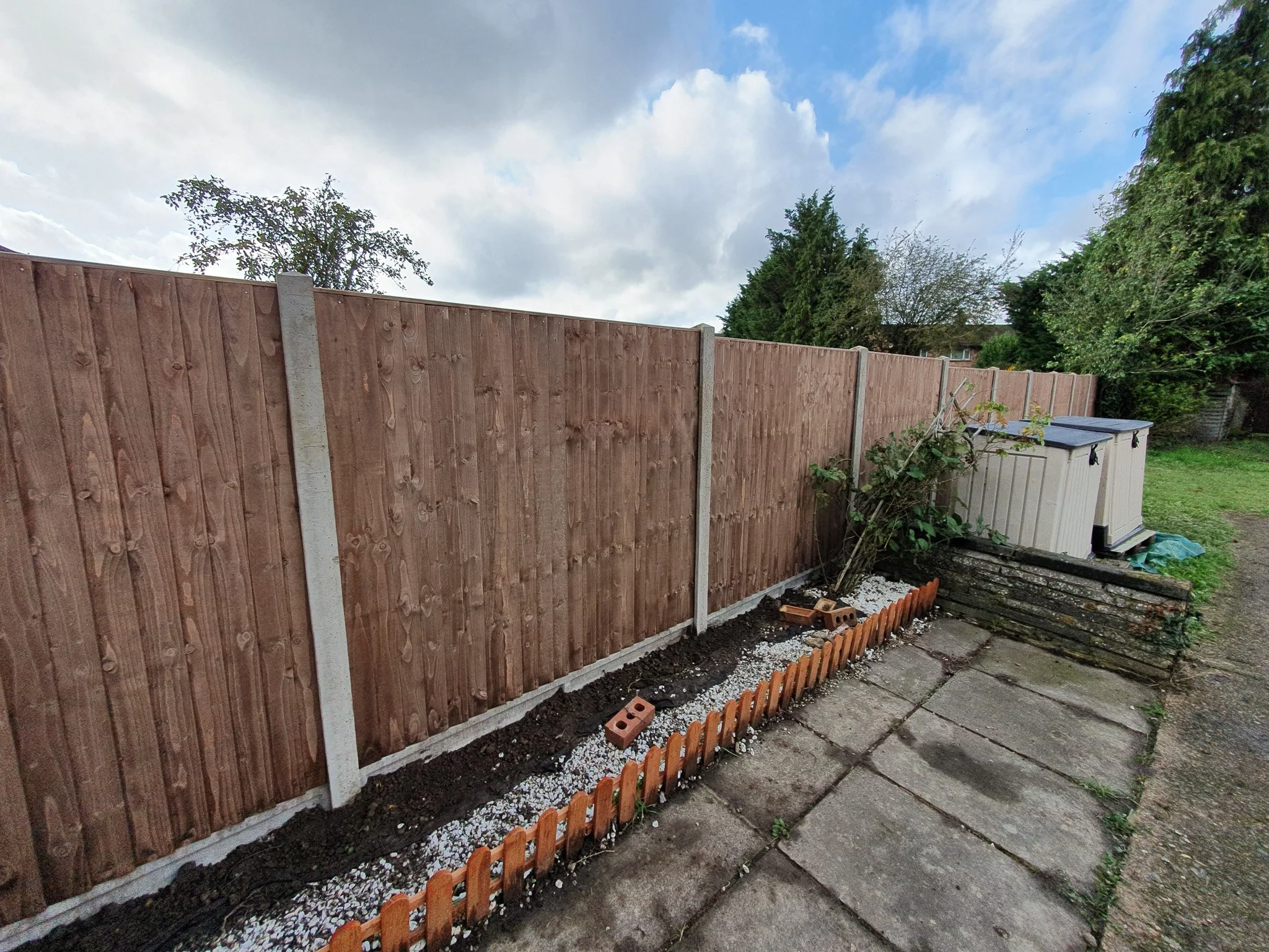 Backyard with newly installed wooden fence, small garden bed with a miniature fence, potted plants, compost bins, and overcast sky