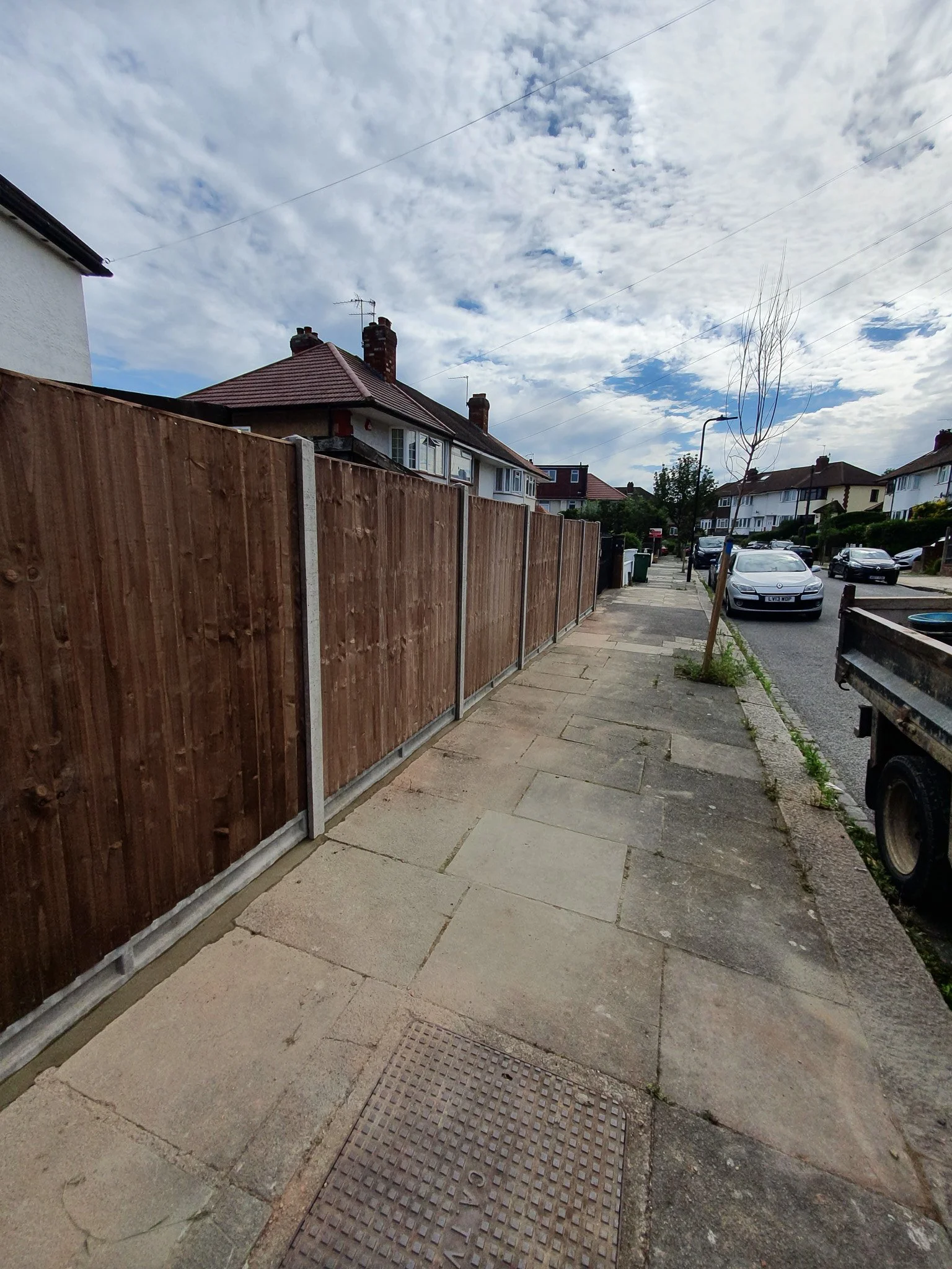 A sidewalk with a wooden fence on the left, a small tree on the right, and houses and parked cars in the background, under a partly cloudy sky.