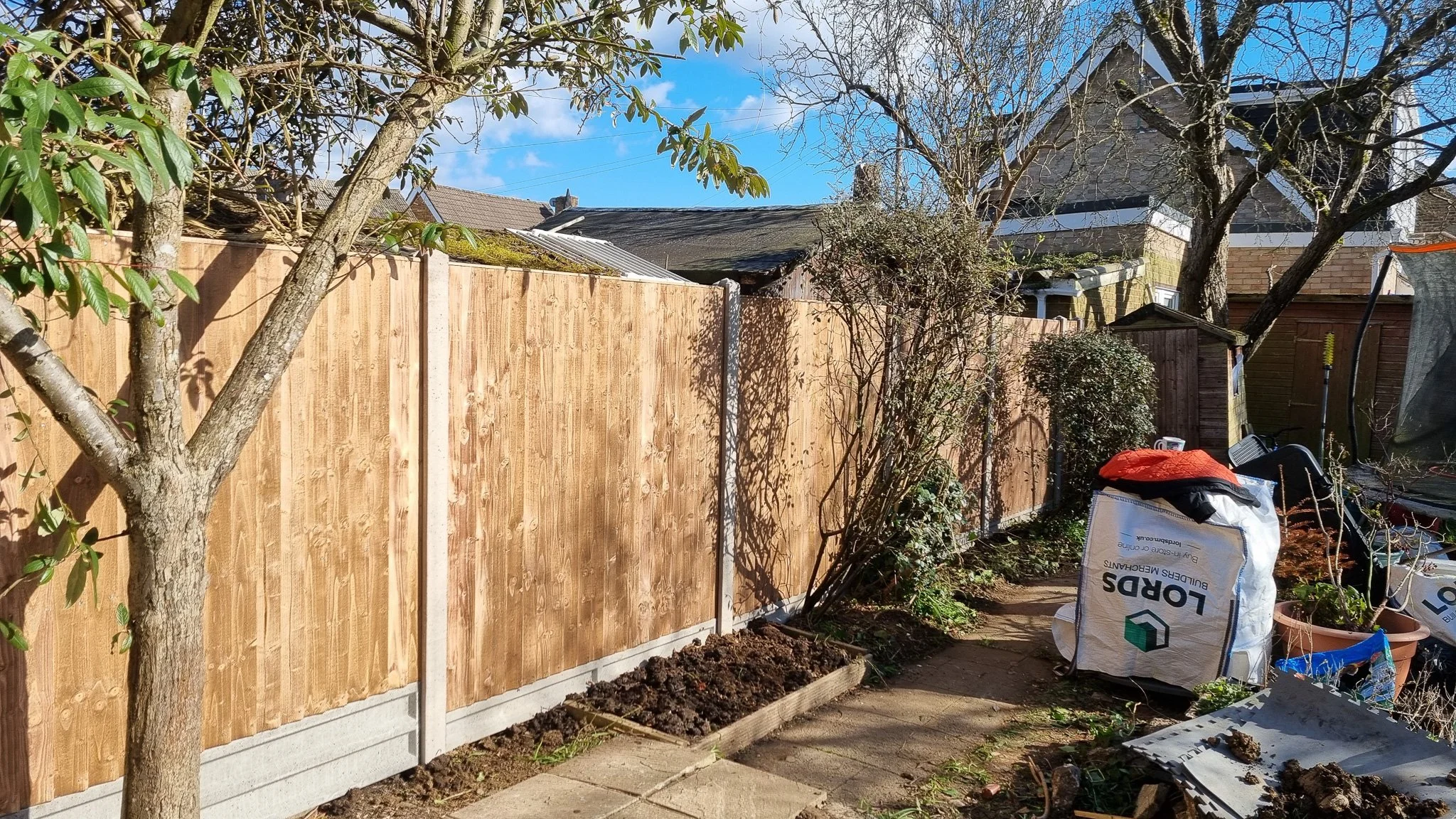 Backyard with newly installed wooden fence, trees, garden tools, bags of soil, and a trampoline.