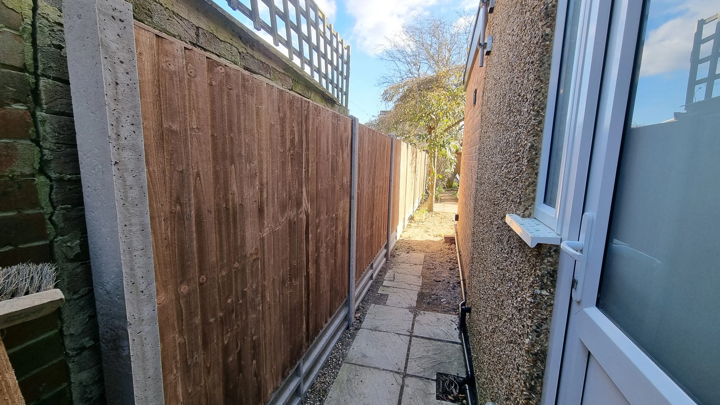 Narrow backyard passage between a brick wall with a window and a newly installed wooden fence, with paving stones on the ground and some garden trees visible in the background.